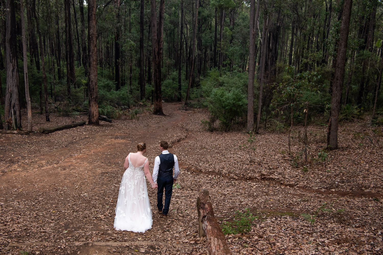 A couple in wedding attire walking down a forest path covered in fallen leaves on their special day.