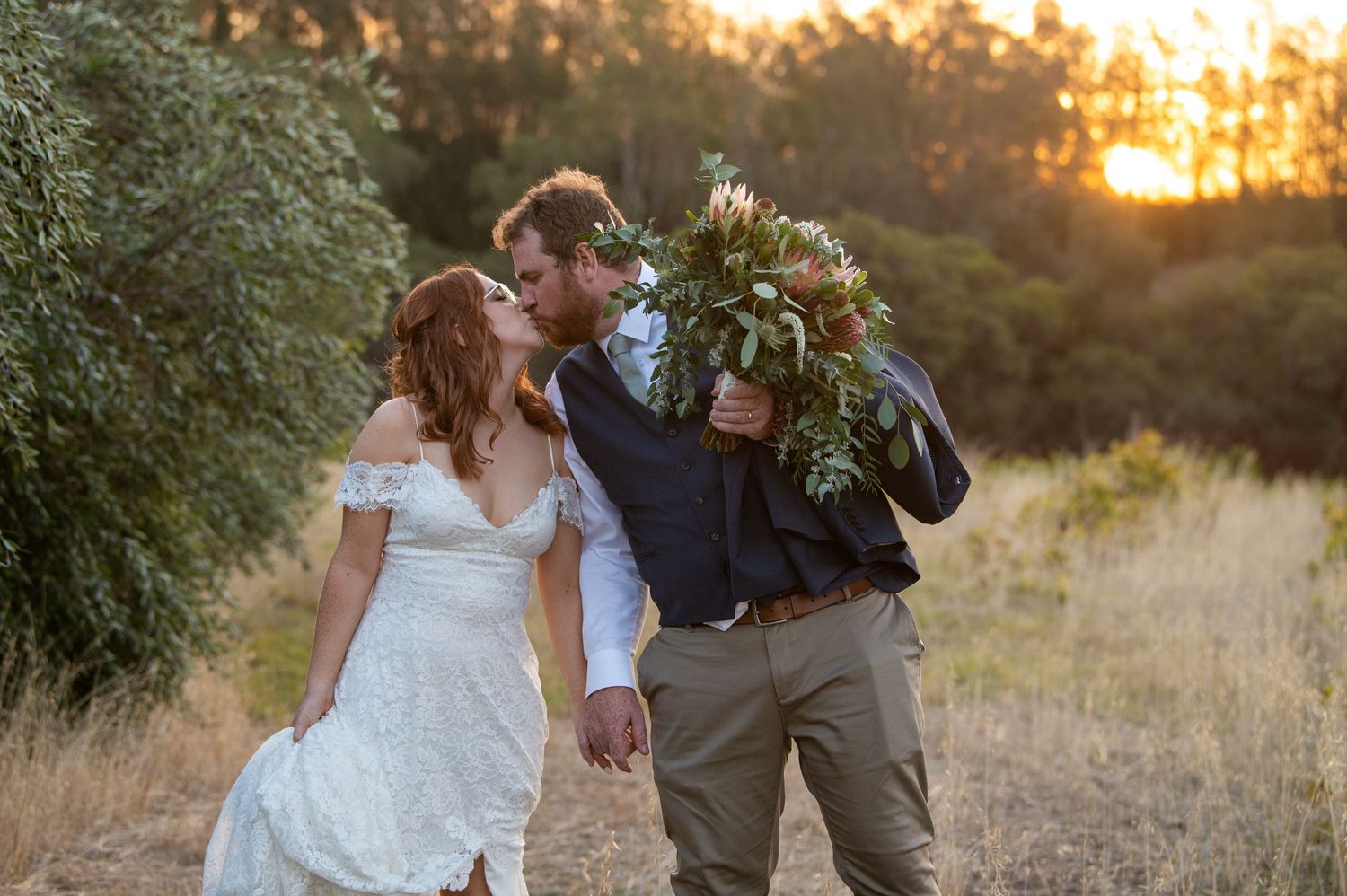 A couple walking and laughing together at sunset while holding a rustic bouquet.