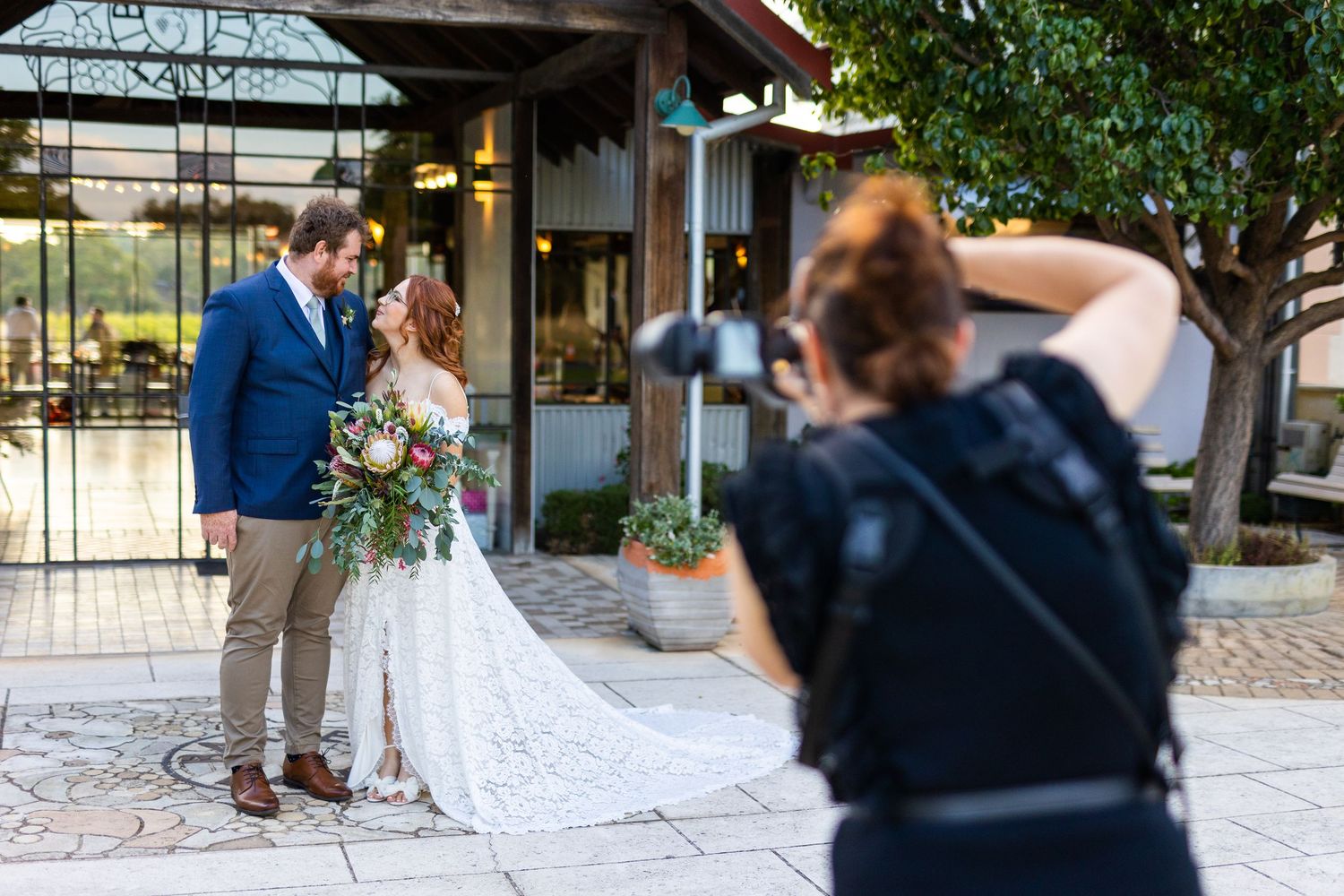 A photographer in black captures a newlywed couple posing together outside a rustic venue with floral bouquet.