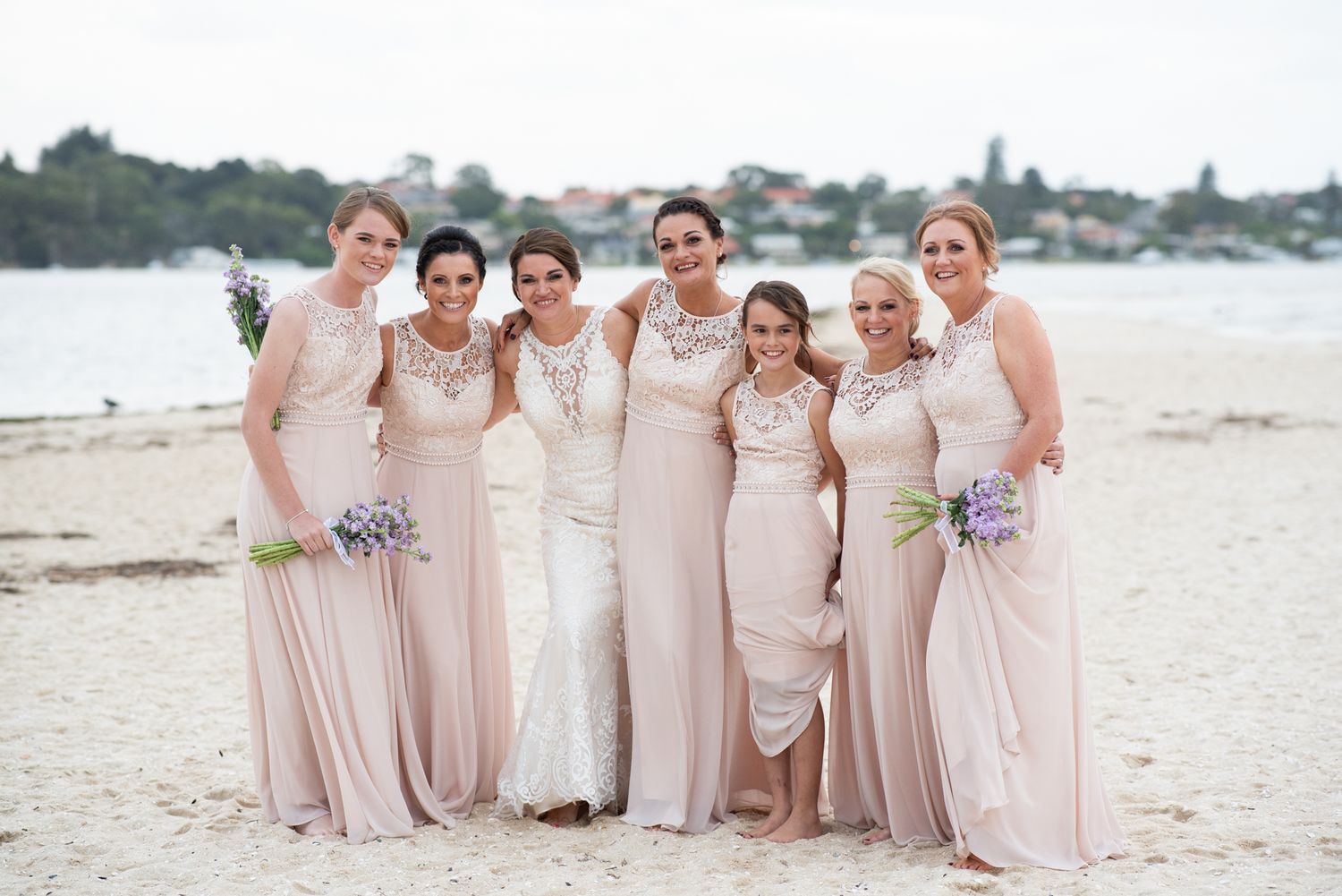 Groupshot of bride with her bridesmaids in blush pink dresses pose together on beach with water and houses in background.