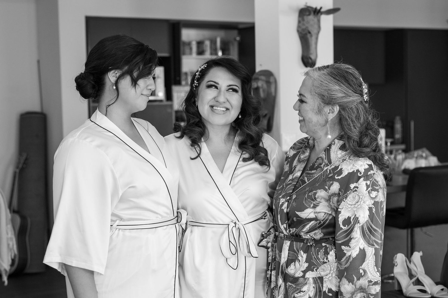 Three people in white robes and floral attire share a joyful moment while getting ready for a special occasion.