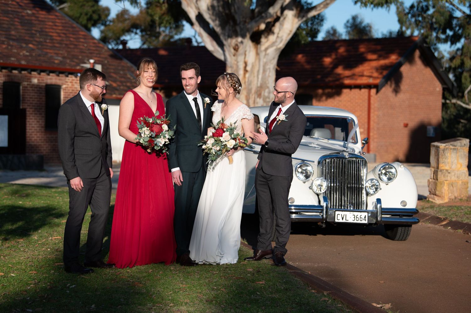 Wedding party gathered outdoors with red and black formal attire against a brick building backdrop.