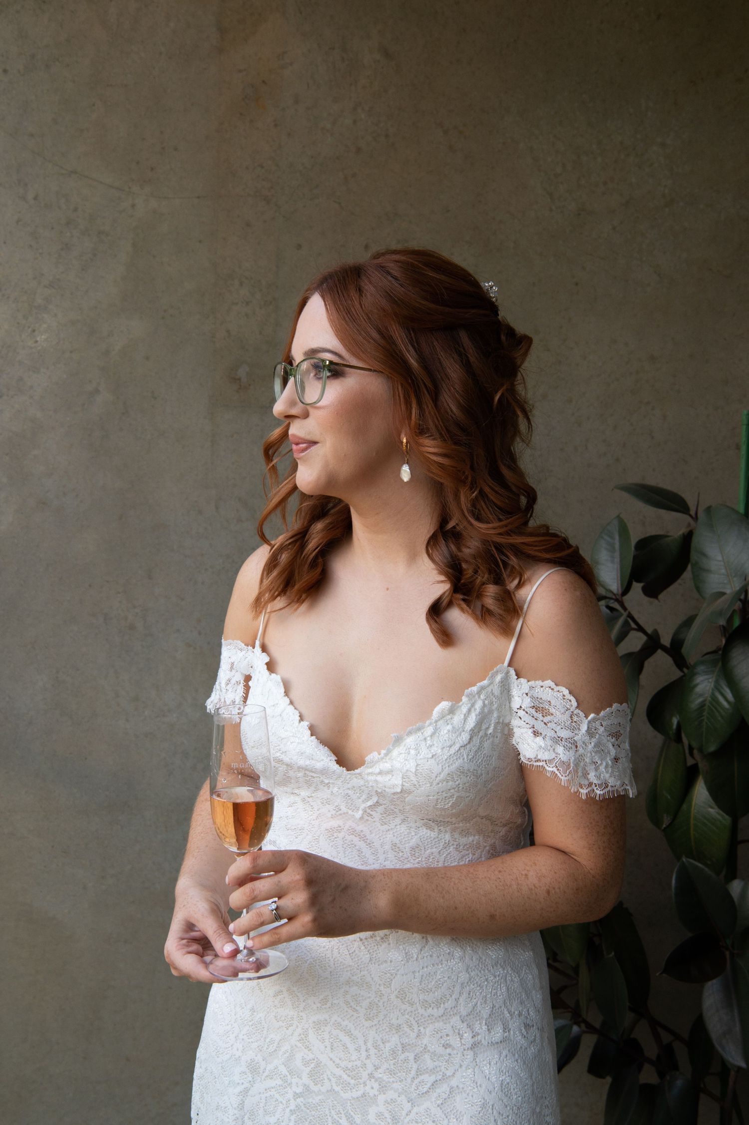Side profile portrait of bride wearing wearing a white lace wedding dress and glasses while holding a champagne flute.