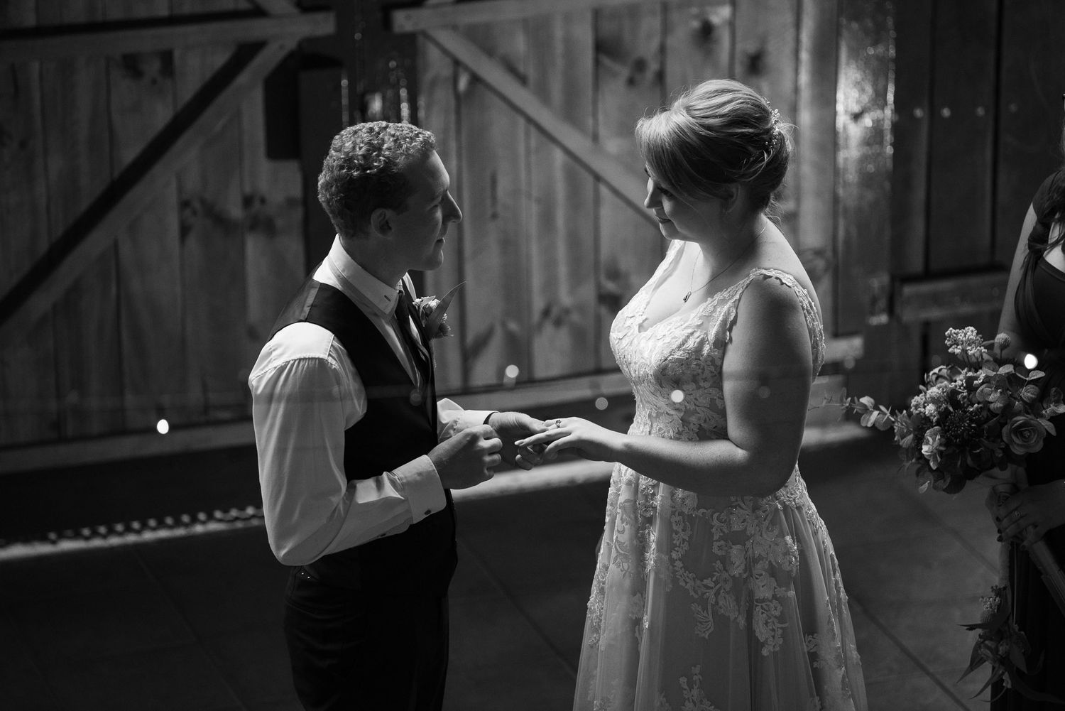 A couple exchanges rings during an intimate black and white wedding ceremony in a rustic barn setting.