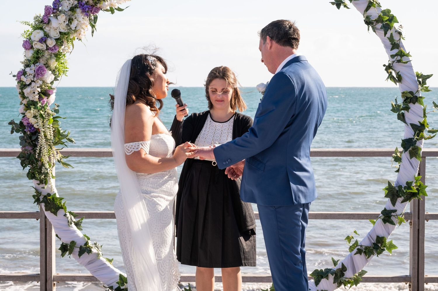 A wedding ceremony takes place under a floral arch by the ocean with gentle waves in the background.
