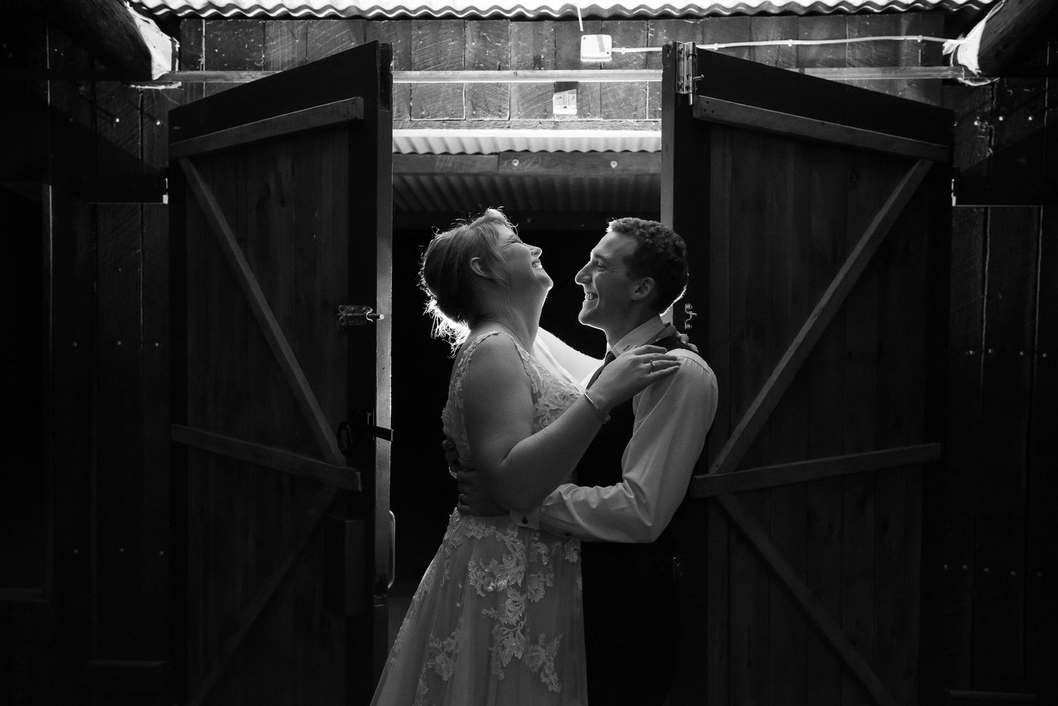 Two people in wedding attire share an intimate dance moment in black and white between rustic barn doors.