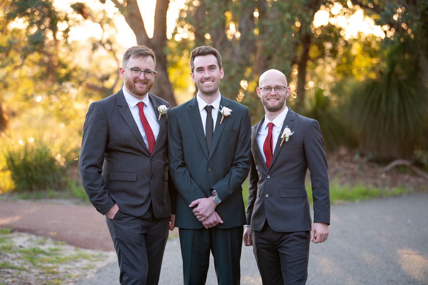 Groomsmen in formal suits pose outdoors together during golden hour at a wedding.