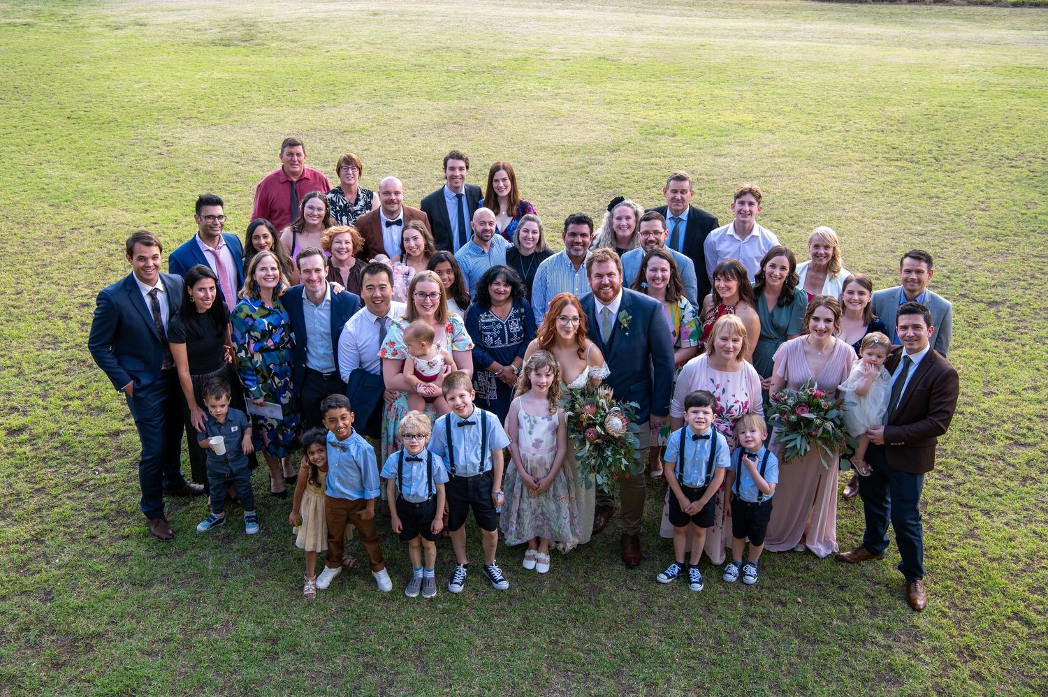 Large group photo of wedding guests gathered outdoors on a grassy lawn.