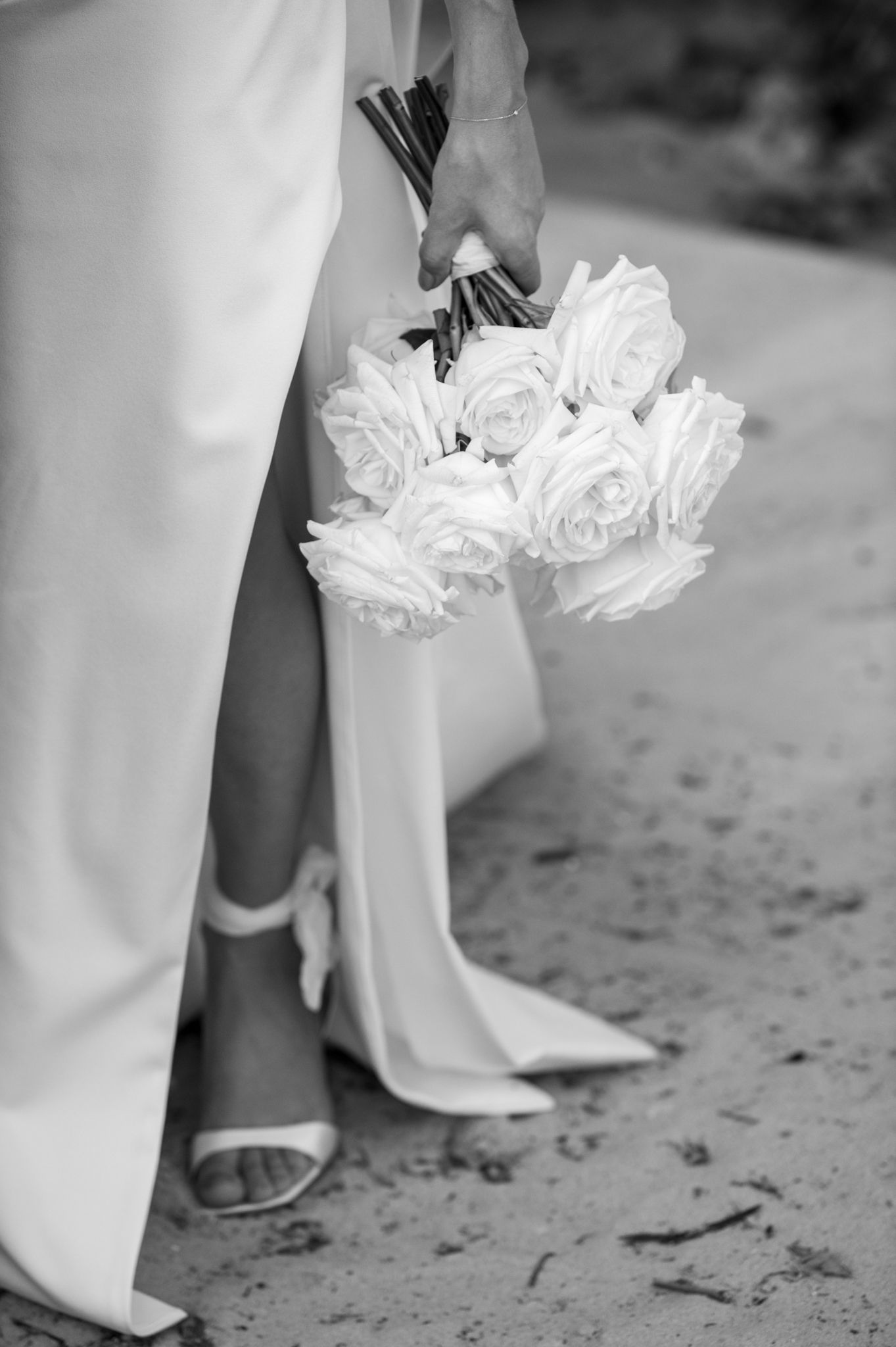 Black and white photograph showing a bride holding bouquet of white roses standing on a sandy ground at a beach ceremony.