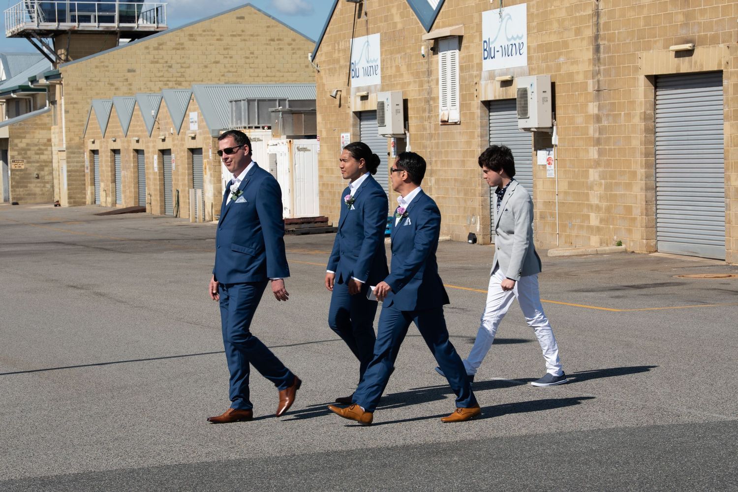 A group walking outside past beige brick buildings wearing navy suits and business attire in bright sunlight.
