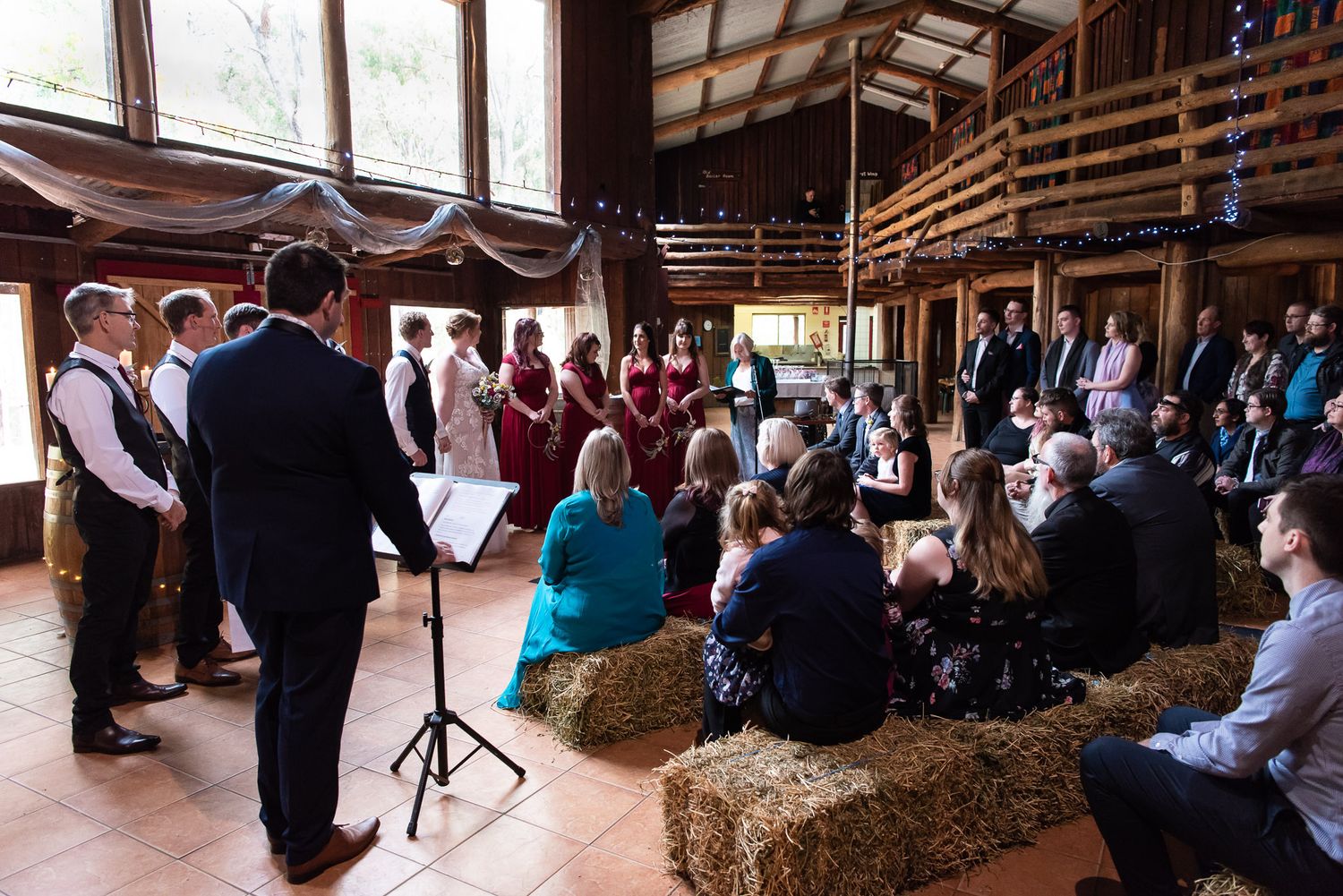 A rustic barn wedding ceremony takes place with guests seated on hay bales as a couple exchanges vows at the front.