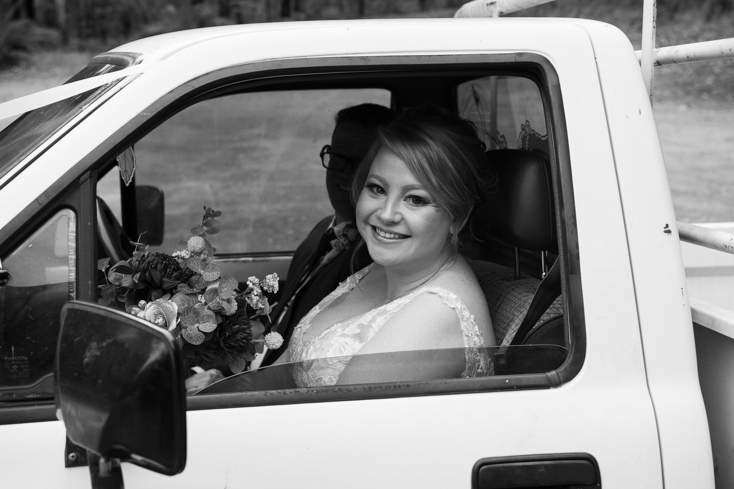 Happy bride smiling through car window with elegant veil and floral bouquet on wedding day.