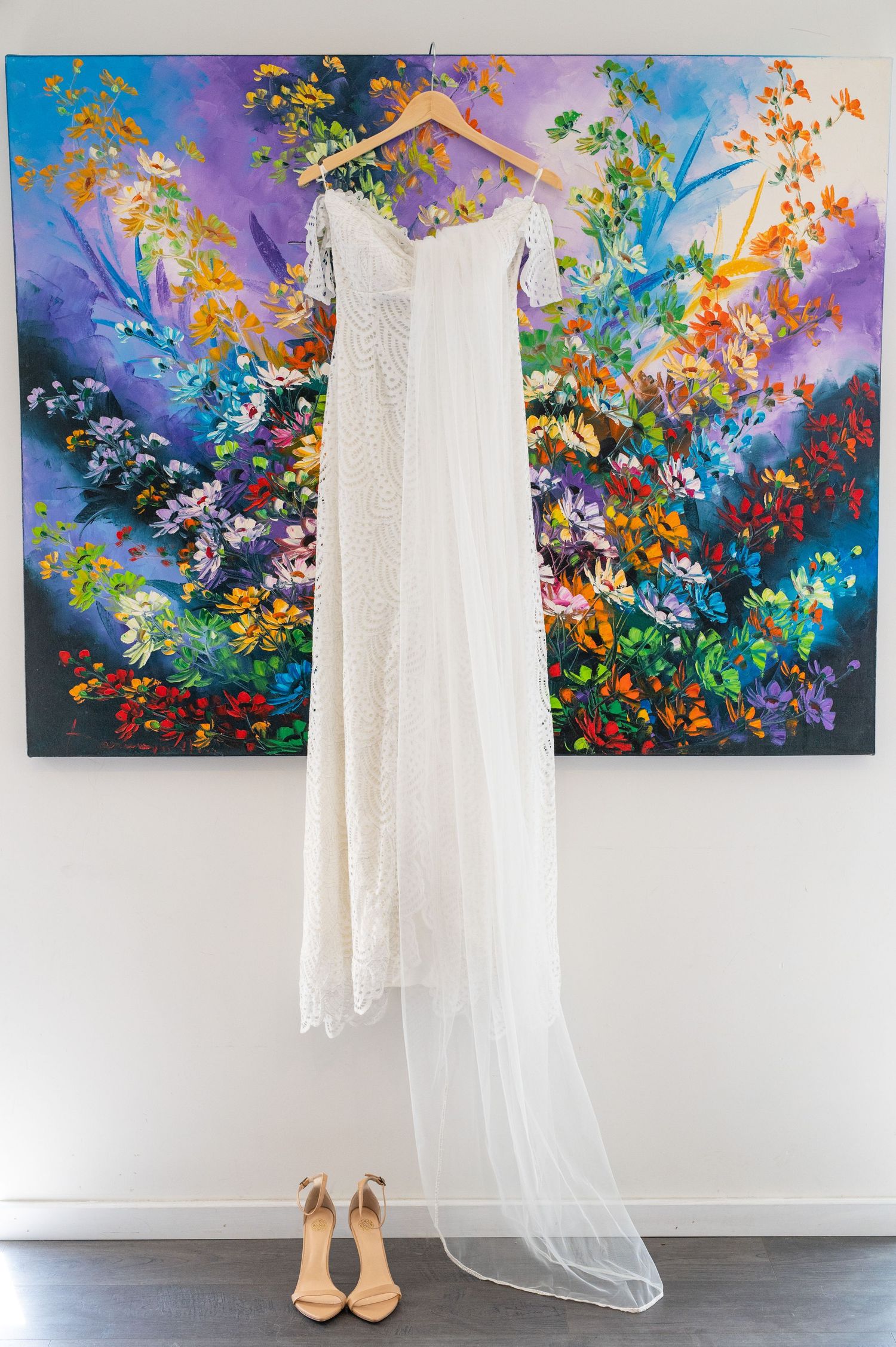 White lace wedding dress hangs elegantly against vibrant floral art with nude heels placed underneath.
