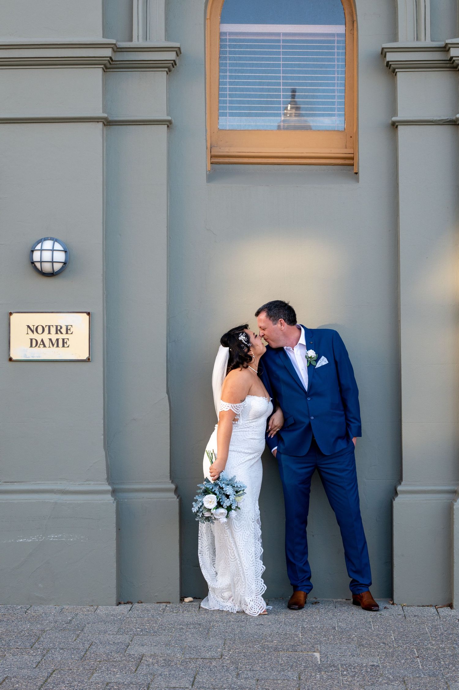 A romantic wedding portrait against a grey wall with a 'Notre Dame' sign and arched window taken in Fremantle, Perth