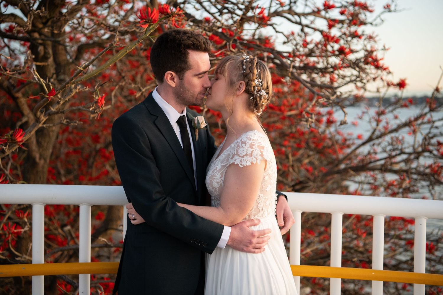 Newlywed couple embracing near a white railing with vibrant red autumn foliage in the background.