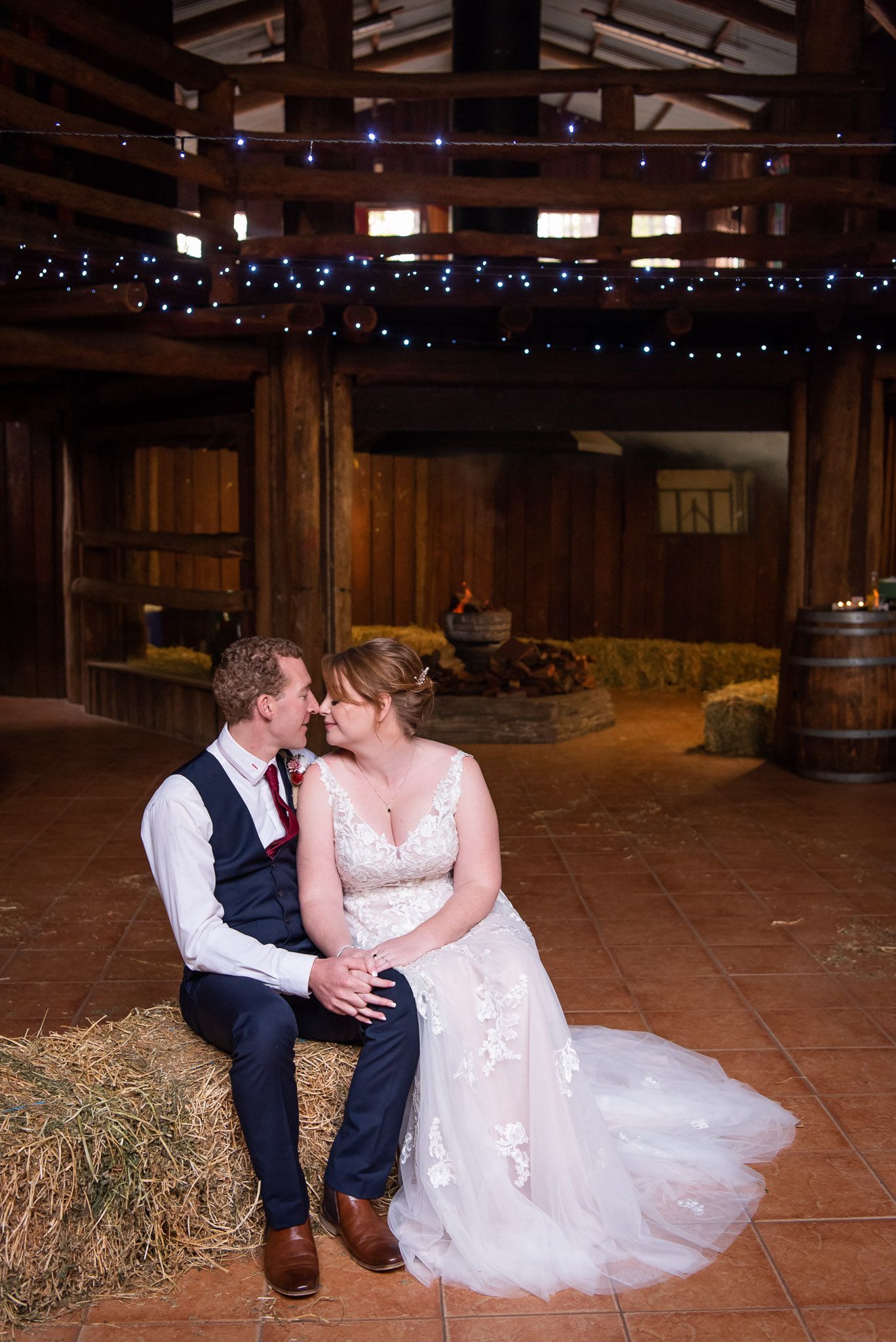 A couple share an intimate moment in a rustic barn venue decorated with twinkling blue lights and hay bales.