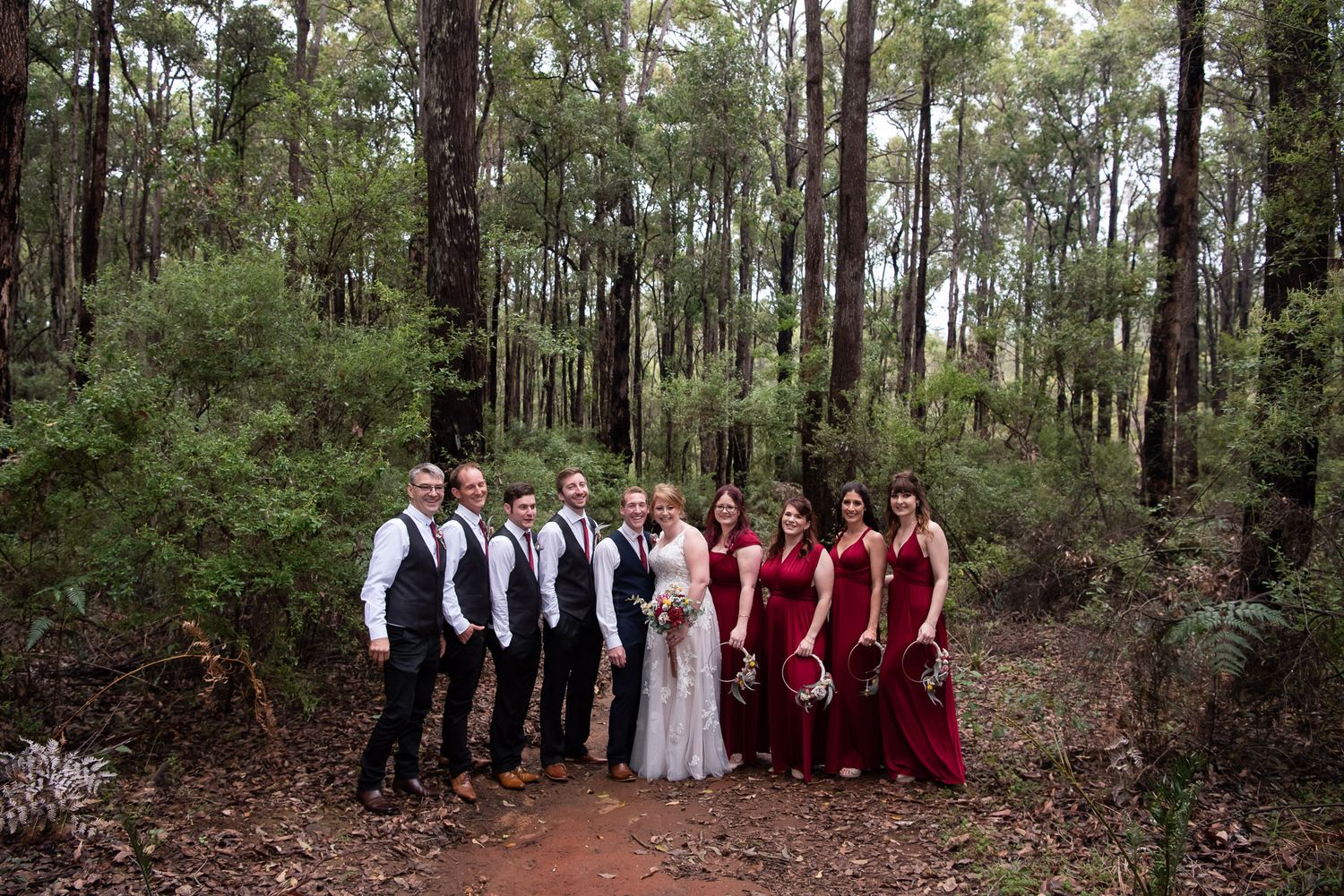 Wedding party in formal attire poses in a scenic forest setting with tall trees and a dirt path.
