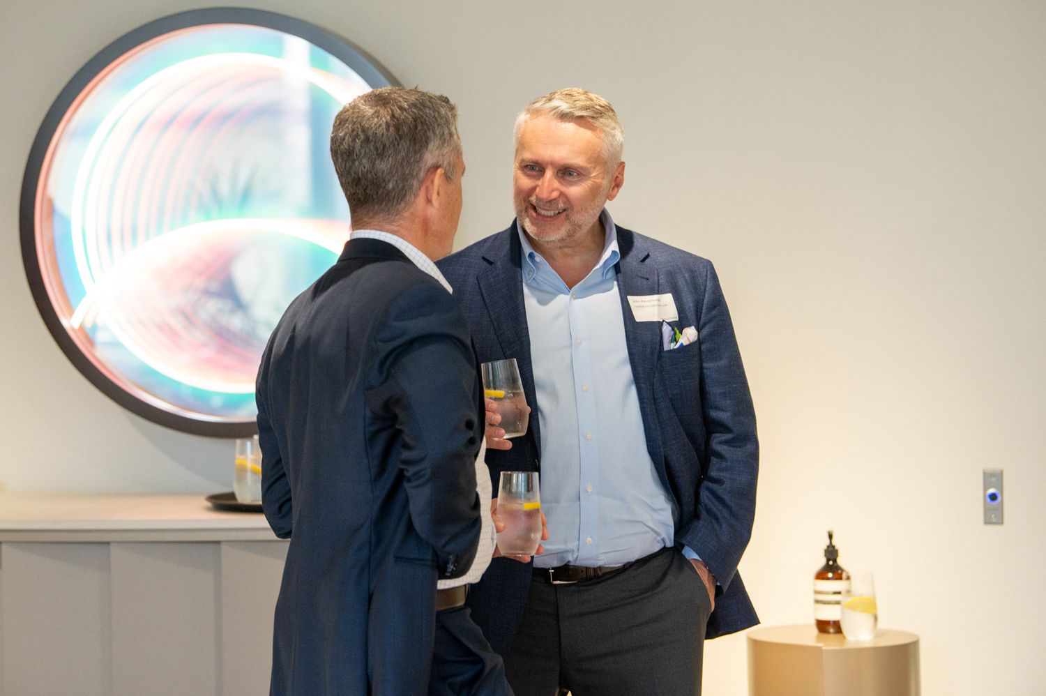 Two professionals in business attire converse and network at a corporate gathering or meeting space.