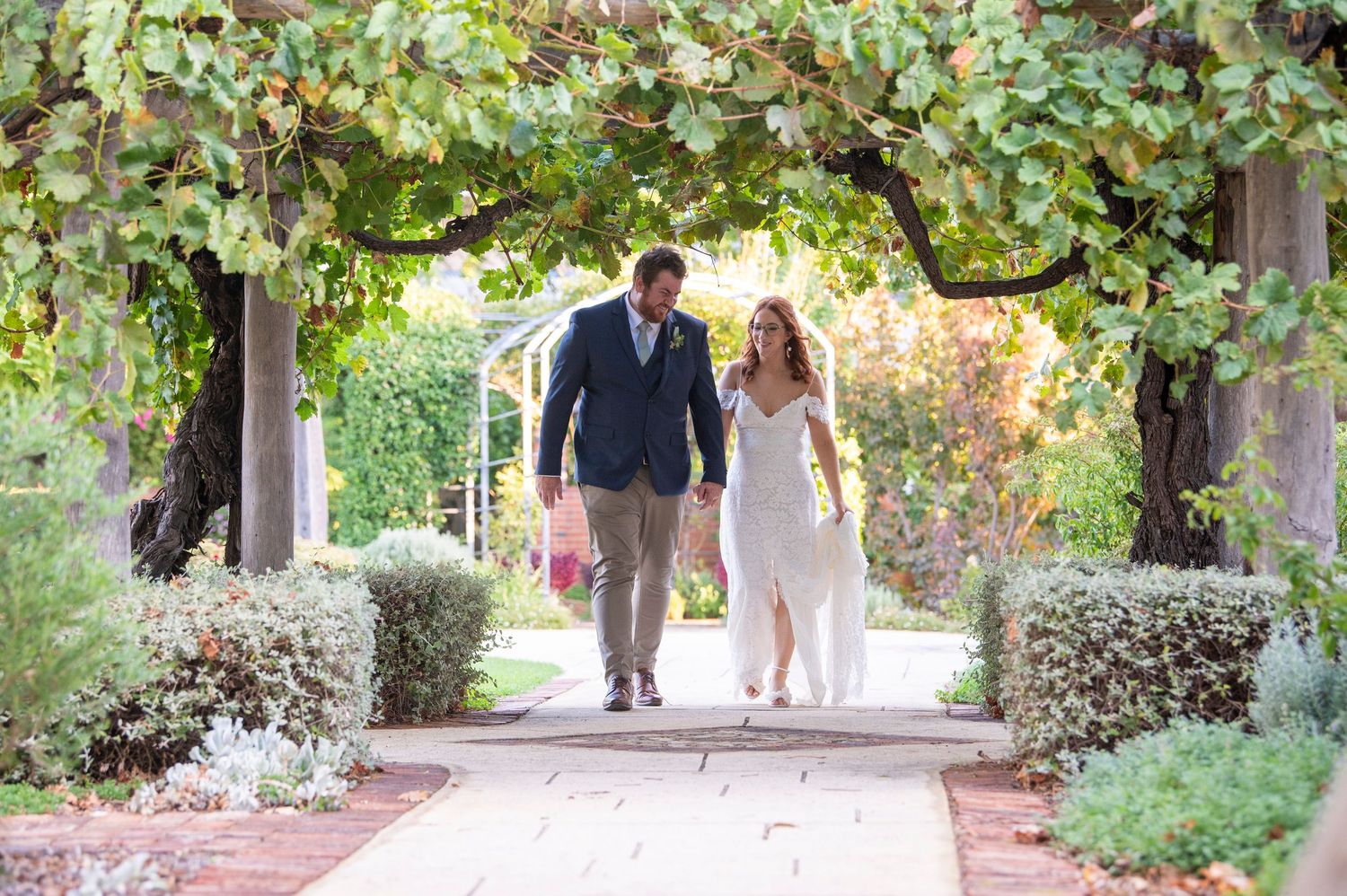 A couple walks down a serene garden pathway surrounded by lush greenery and hanging vines in natural daylight.