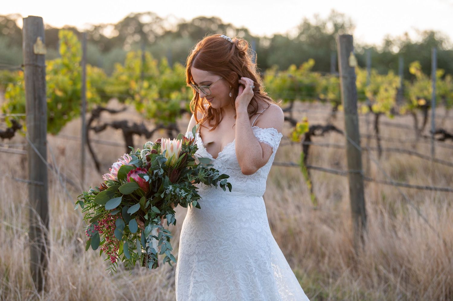A bride in a white lace dress holds a colorful bouquet while standing in a rustic vineyard at sunset.
