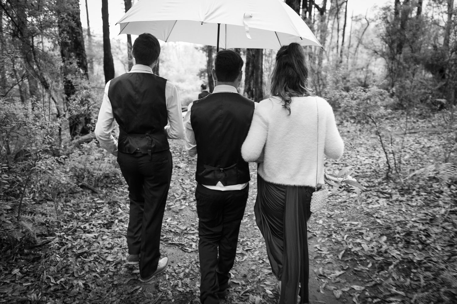 Three people walking together while sharing a white umbrella in a black and white outdoor scene.