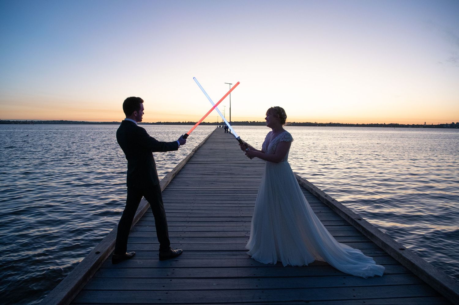 Wedding couple and party posing at sunset on a wooden pier extending over calm waters.