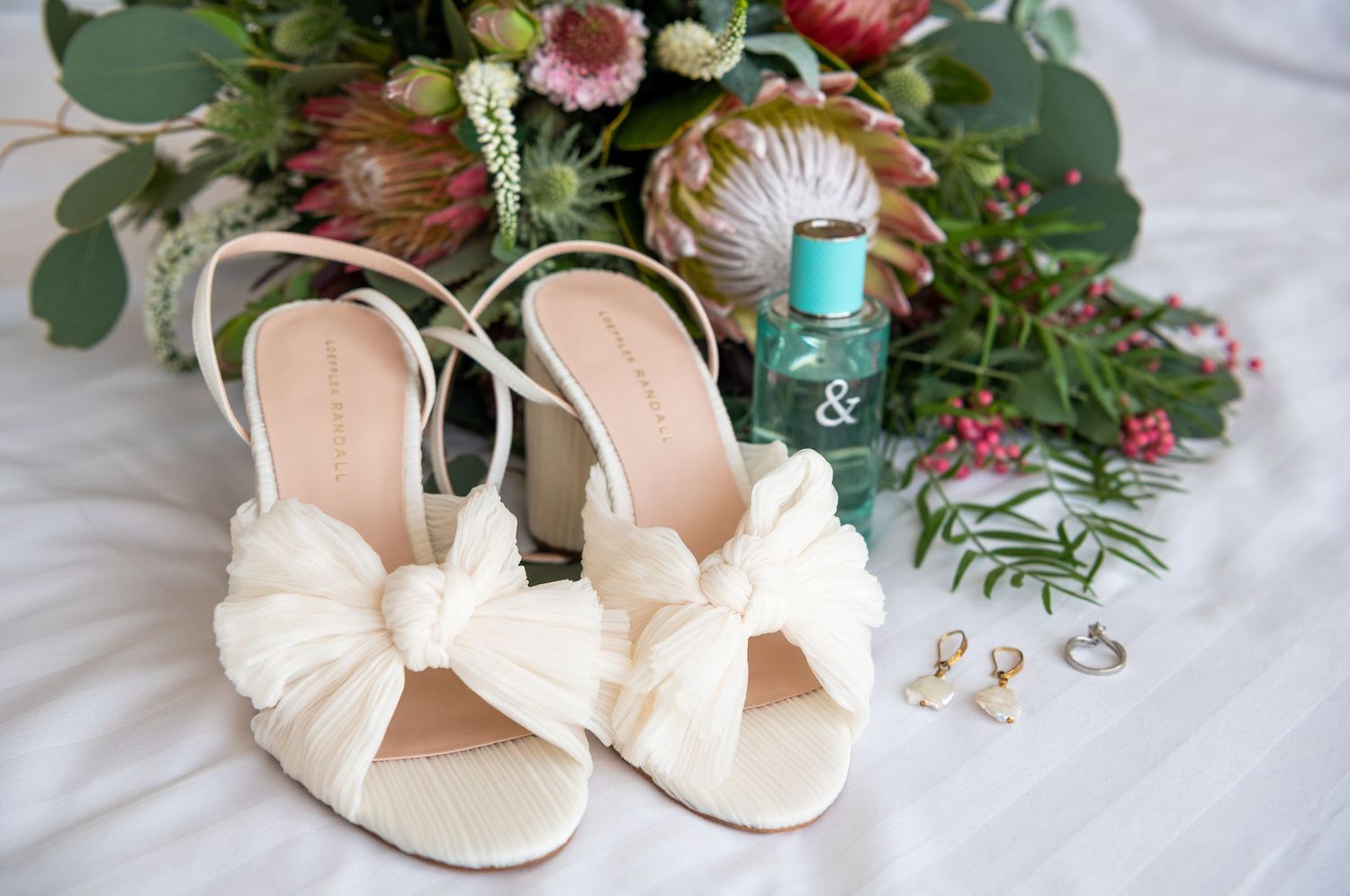 Wedding accessories including ivory bow heels, rings, and floral bouquet arranged on white fabric.
