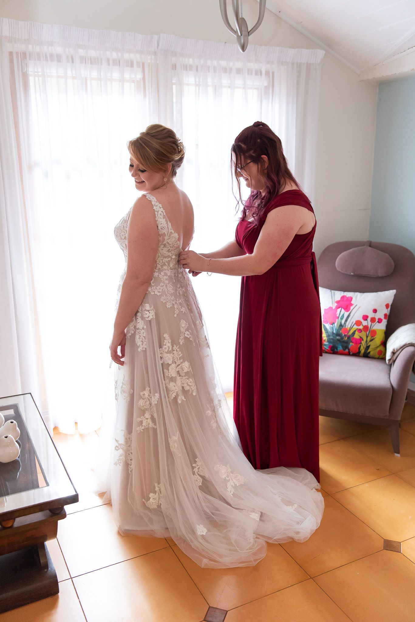 A woman in a burgundy dress helps another in an elegant lace wedding gown with final preparations in a sunlit room.