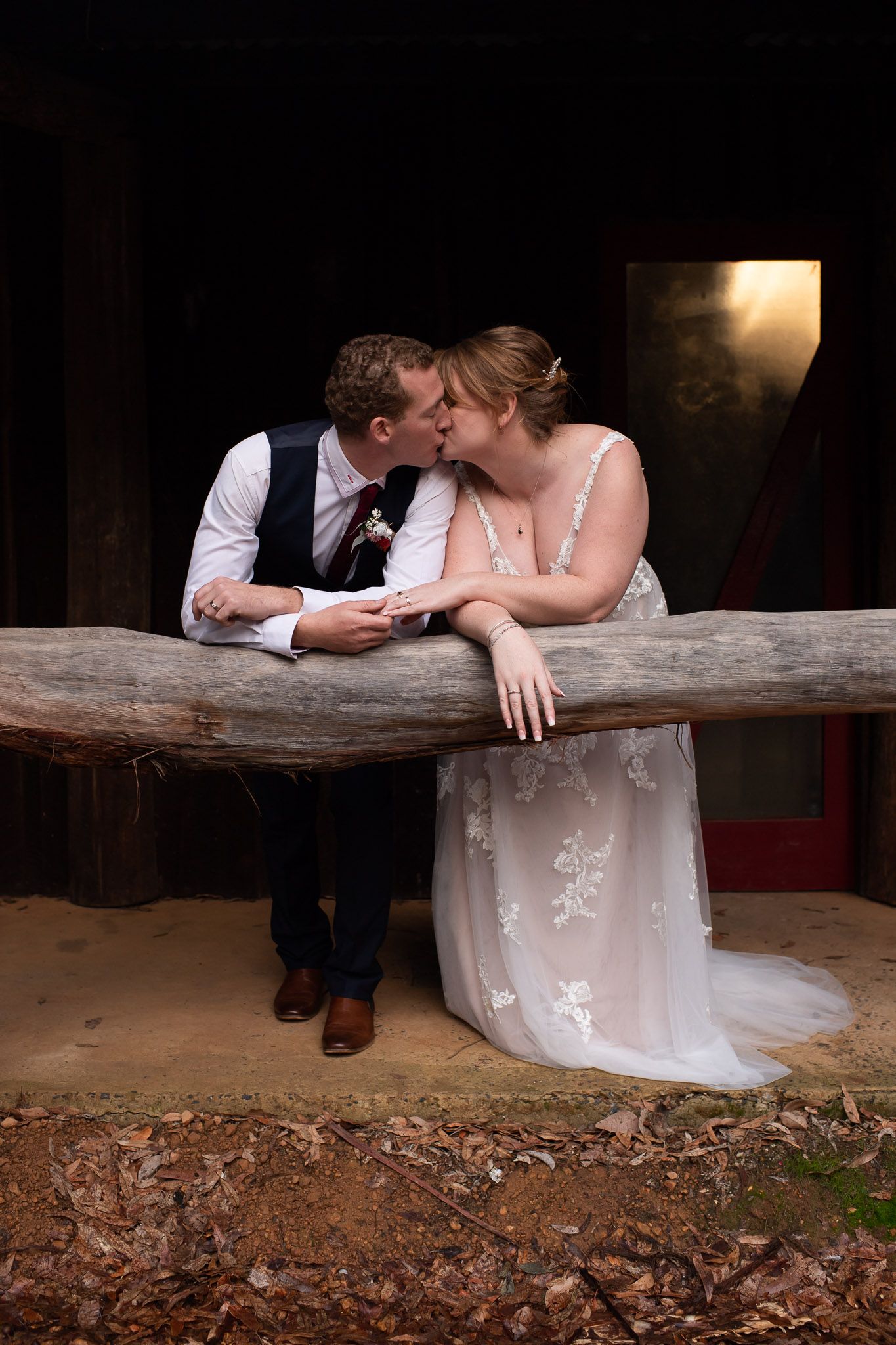 A romantic couple shares a tender moment on a rustic wooden bench at dusk, the bride wearing a delicate lace gown.