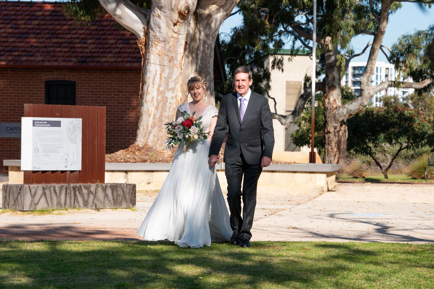 Wedding party walks along a tree-lined path at an outdoor venue during a sunny afternoon celebration.