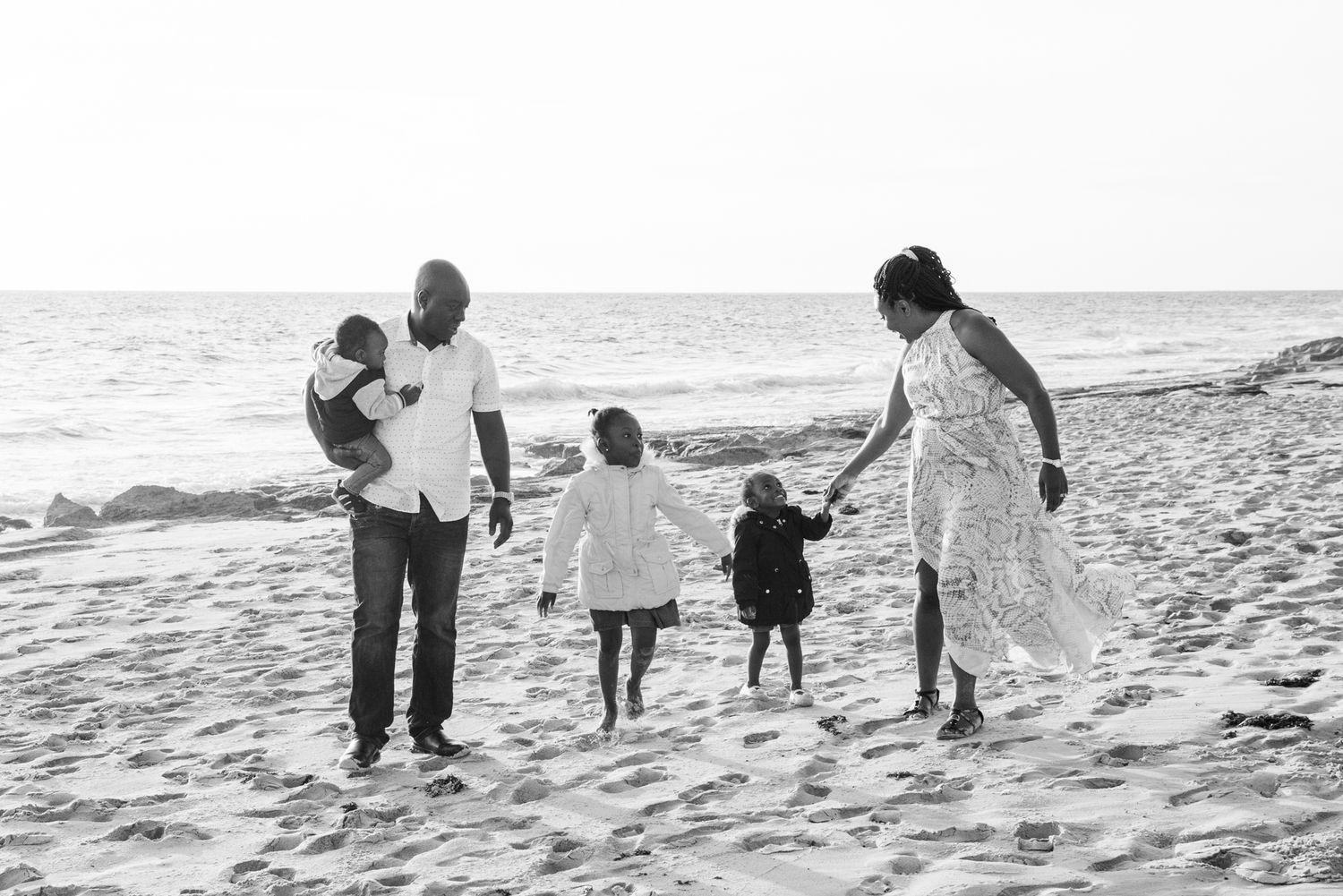 Family walks along the white sandy beach with ocean waves crashing on shore in black and white photography style.