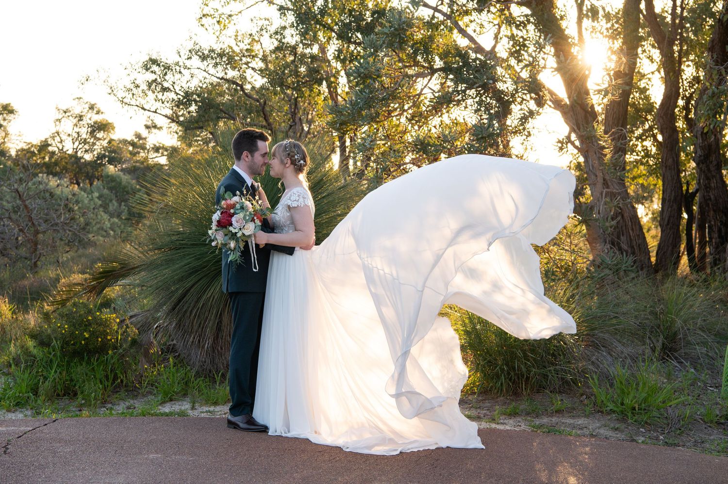 A bride's white dress veil flows dramatically in the wind during a sunset photoshoot among trees.