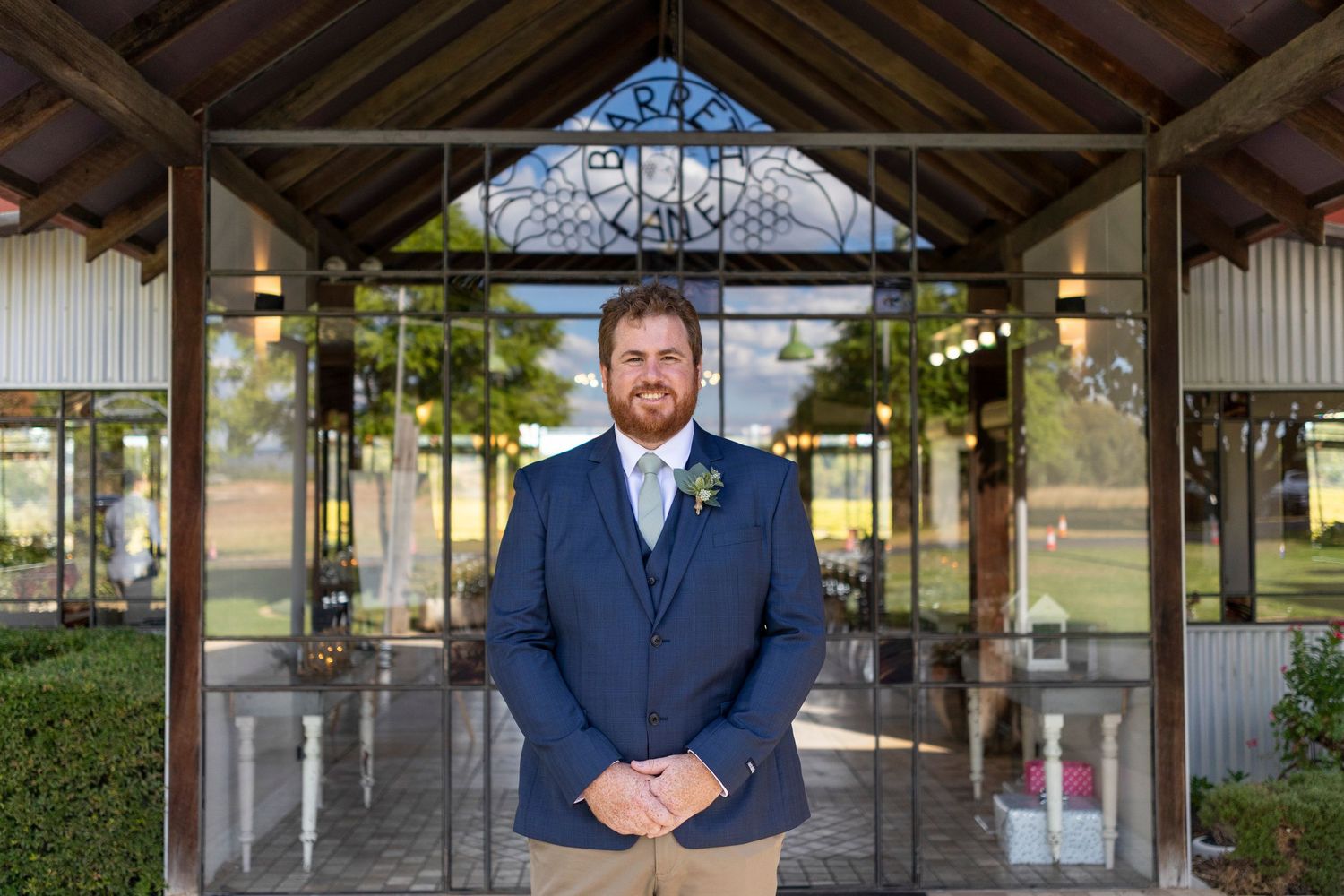 A groom in a navy suit stands in front of a rustic covered outdoor venue entrance with wooden beams.