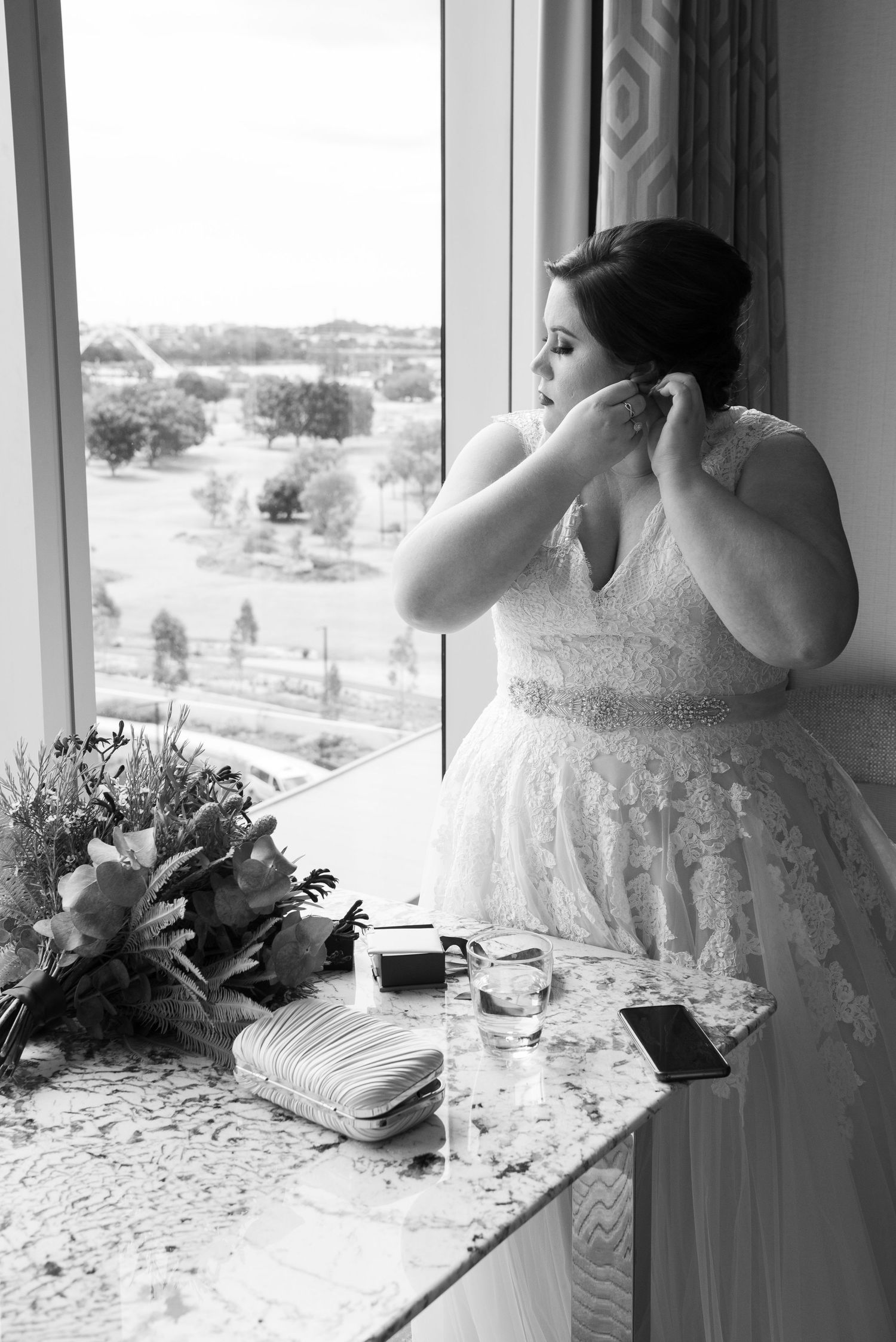A bride in a lace wedding dress puts on earrings while standing at a window, with a bouquet on the counter beside her at Crown Towers, Perth