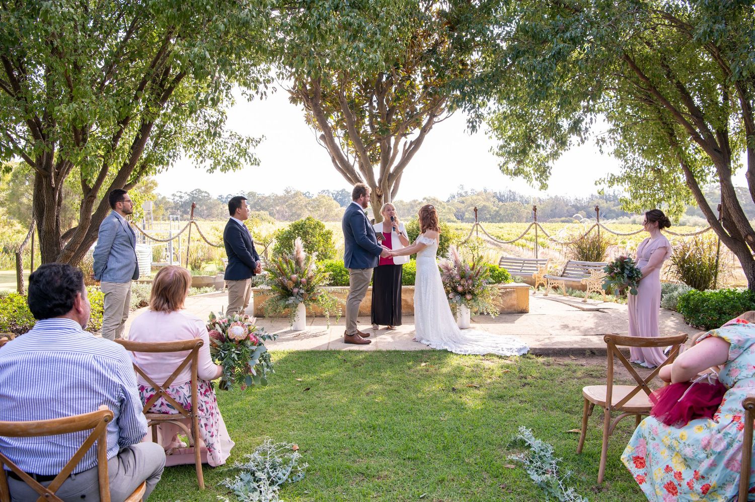Wedding guests seated outdoors under trees on white chairs during a daytime ceremony.