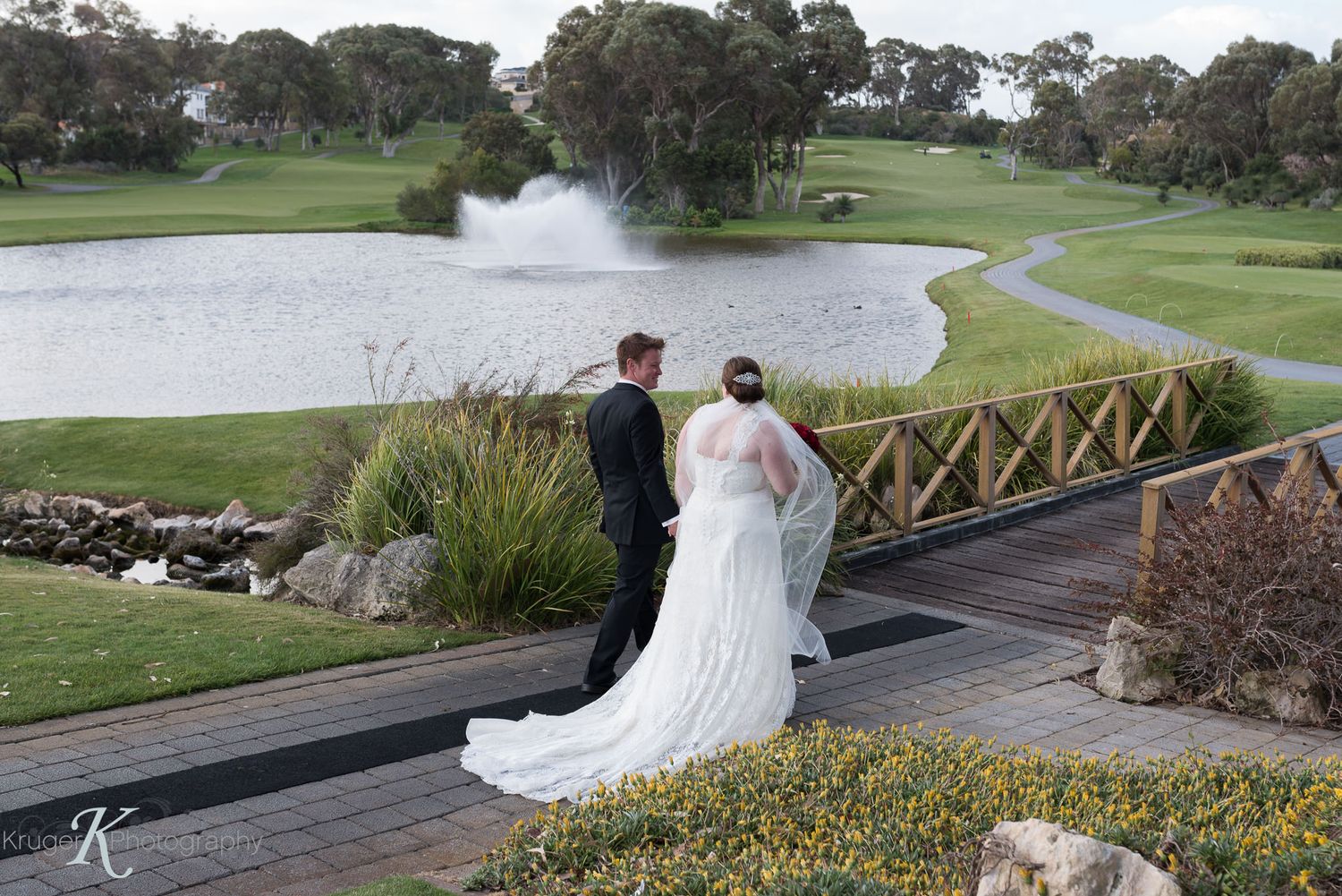 A wedding couple walks over a wooden bridge overlooking a golf course pond with a water fountain.