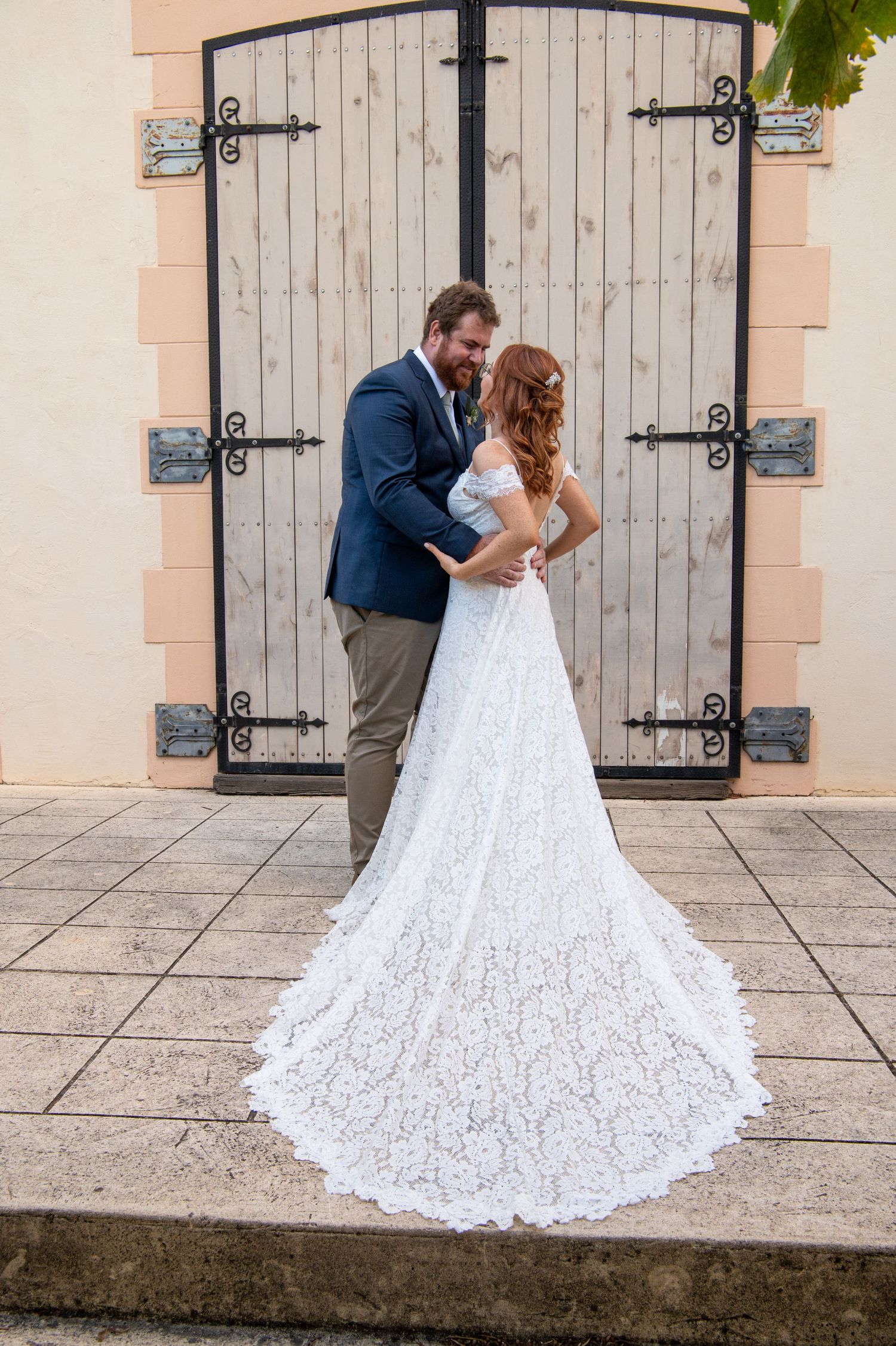 A wedding couple poses against rustic wooden doors with an elegant lace train extending behind.