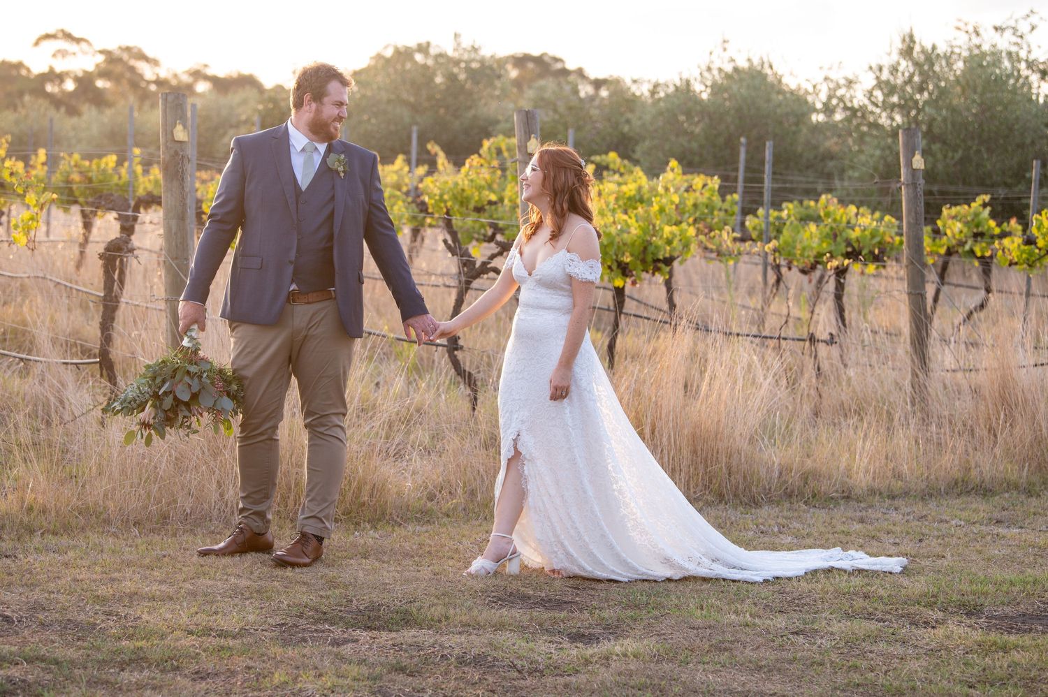A joyful couple walking together through a golden vineyard at sunset with trailing wedding dress details.