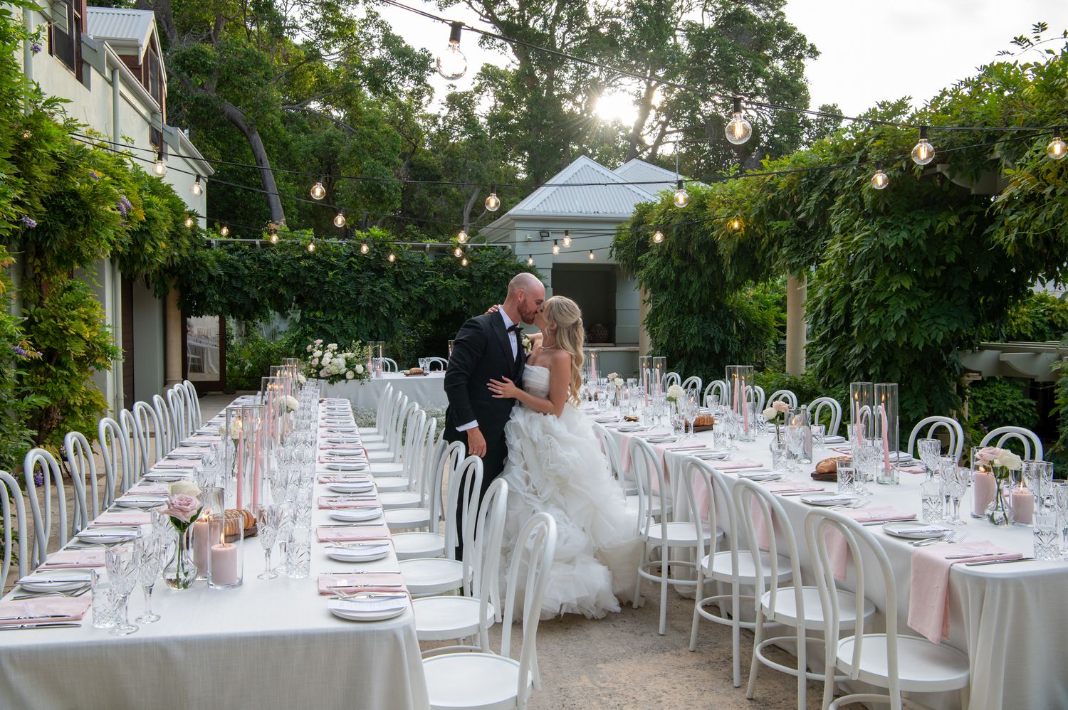 Elegant outdoor wedding reception setup with long white tables, string lights and lush greenery in a garden setting.