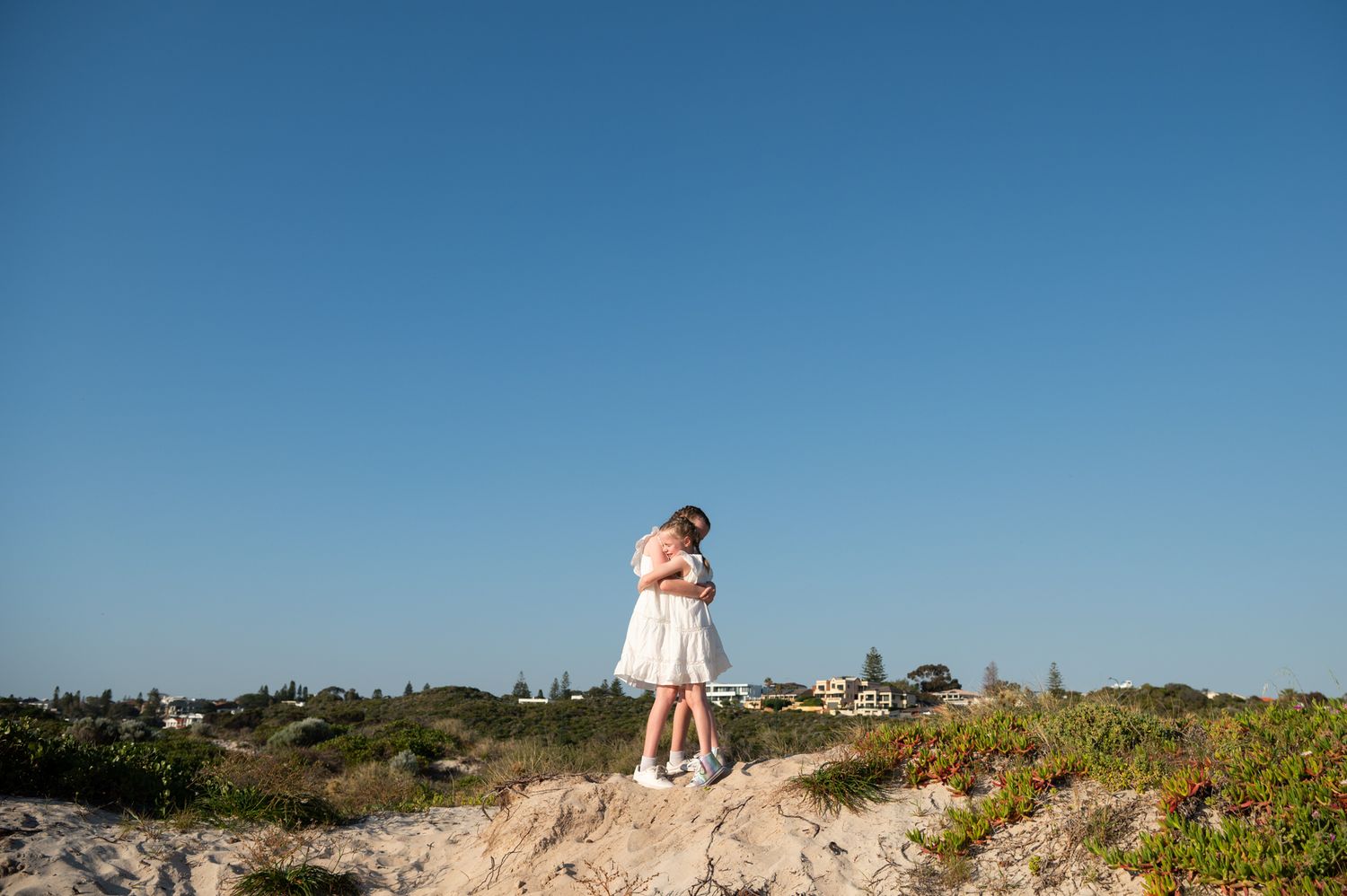 A couple embraces while standing on a sandy dune against a bright blue sky on a sunny day.