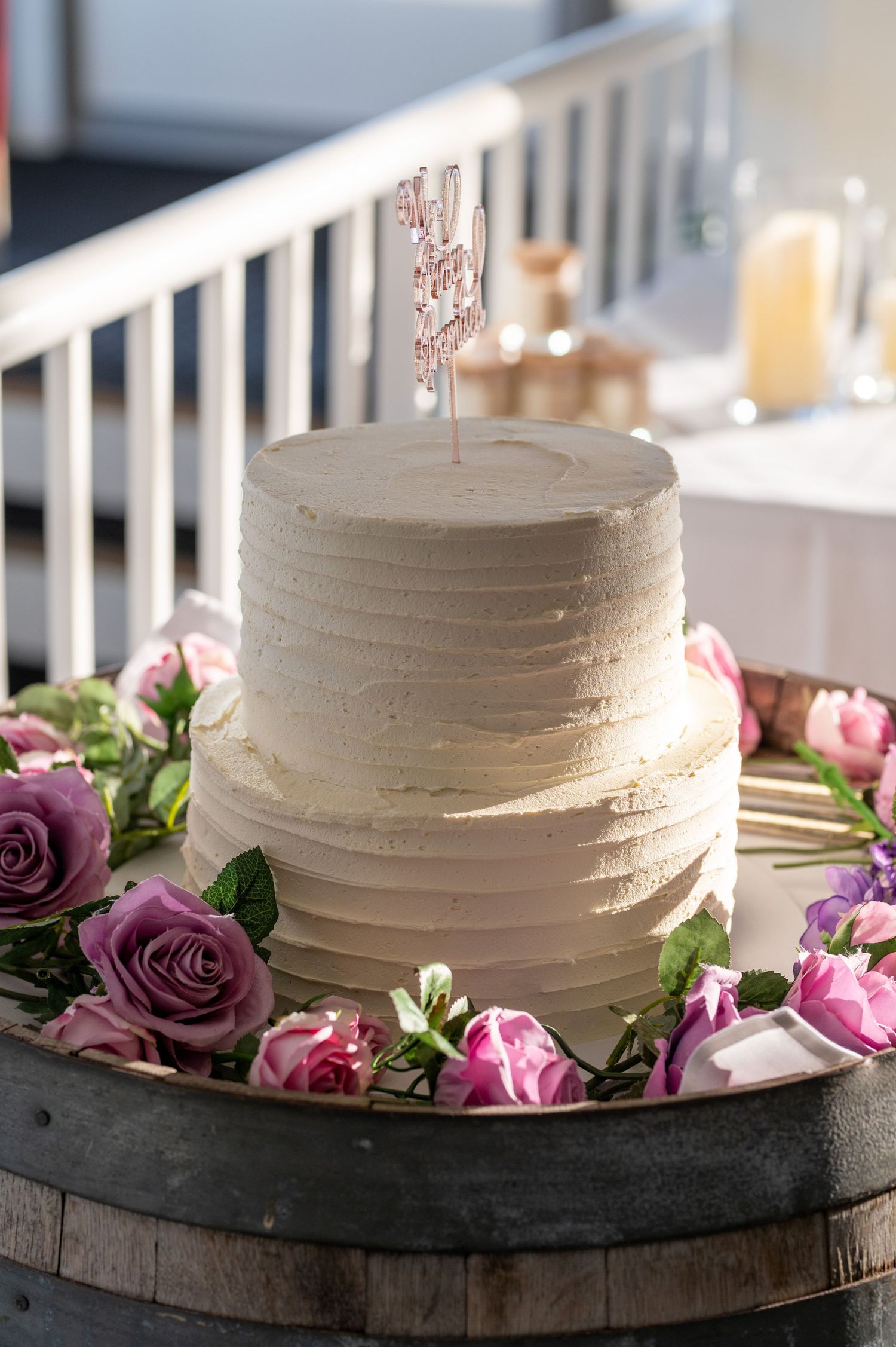 A classic white two-tier wedding cake decorated with pink and purple roses on a rustic wooden base.