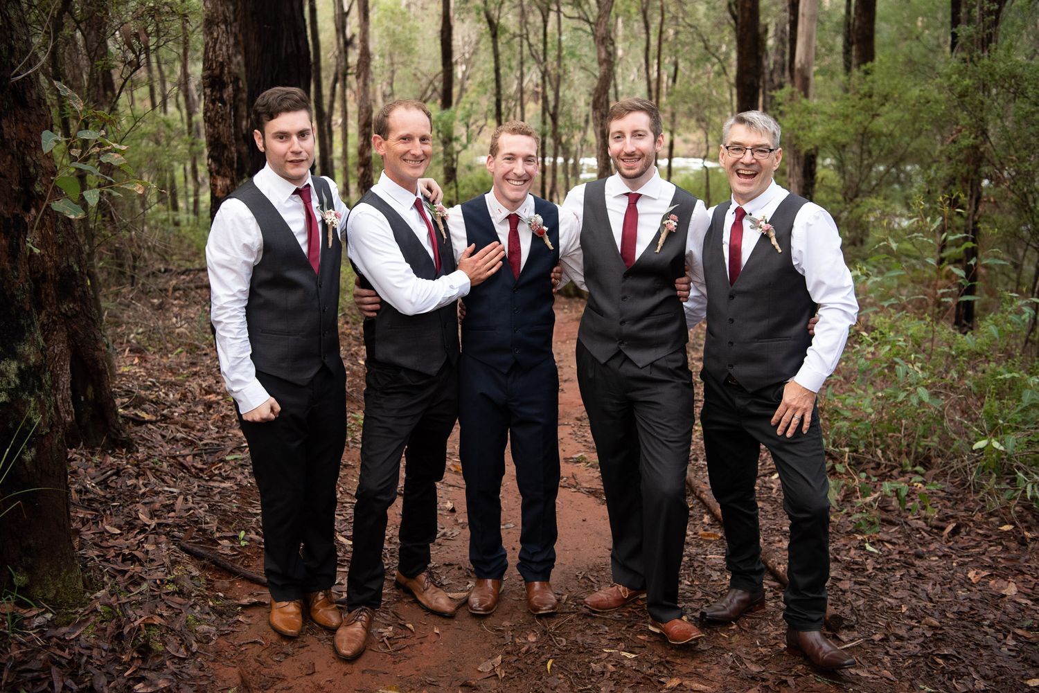 A group of groomsmen in matching black vests and burgundy ties pose together on a wooded forest path.