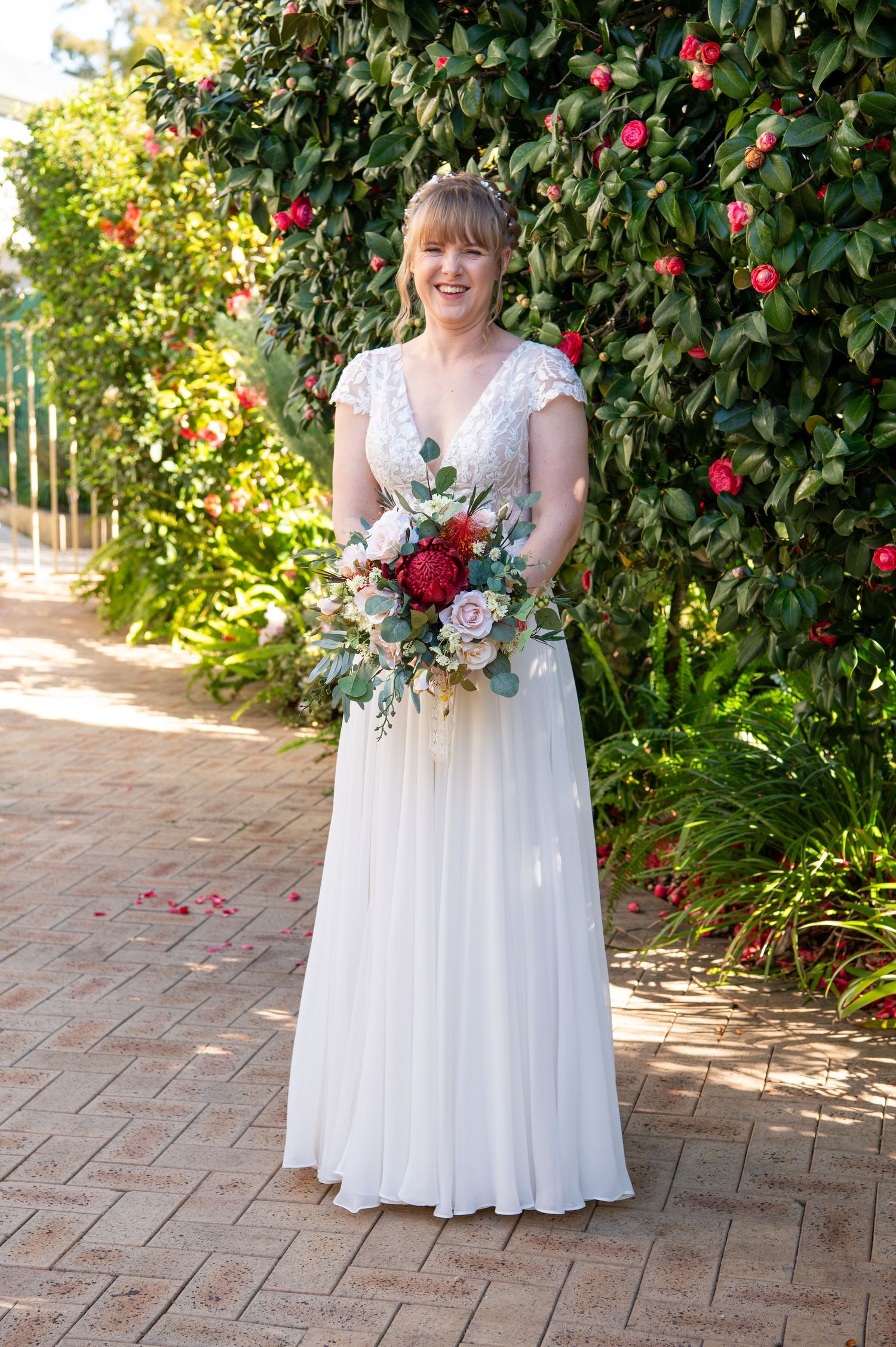 White bridal bouquet with delicate flowers against a backdrop of blooming red camellia bushes and brick pathway.