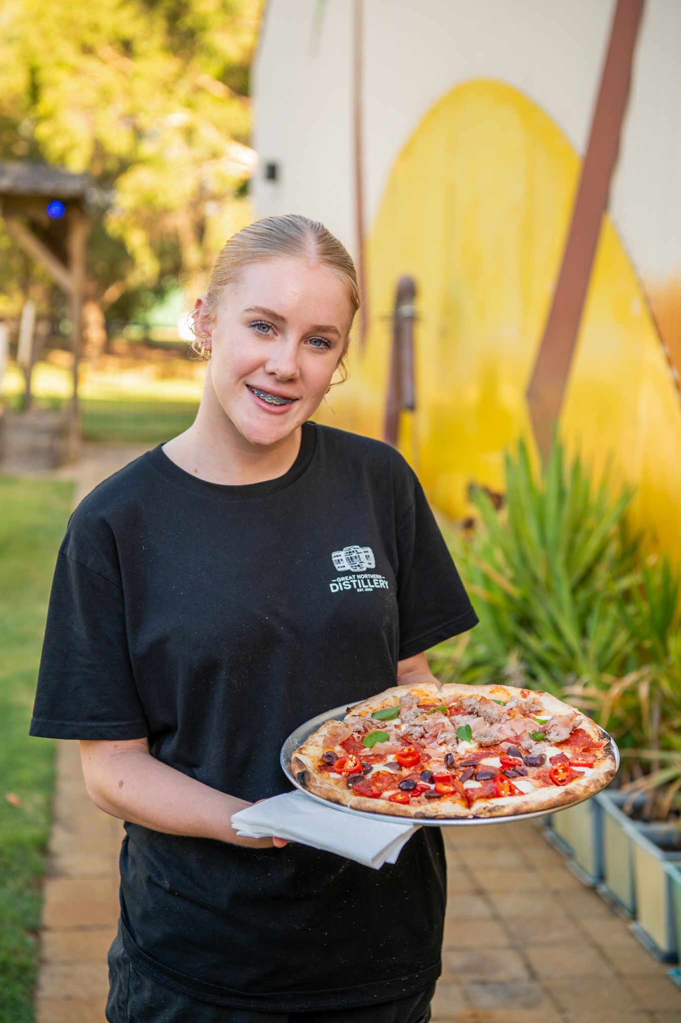 Restaurant staff holding a freshly made pizza with toppings in front of a yellow wall outdoors.