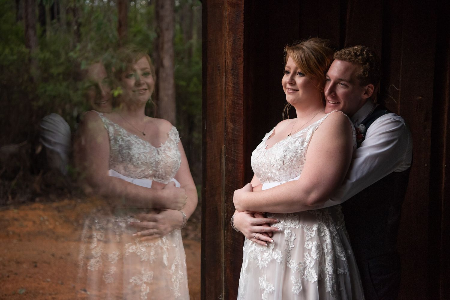 Romantic couple in white wedding attire embrace next to window with reflection on dark wooden frame.