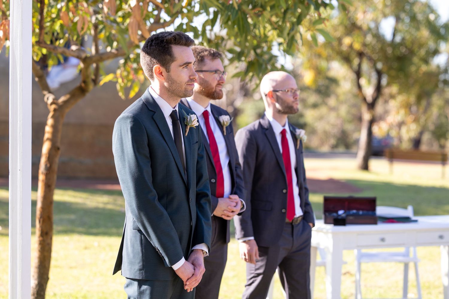 Three groomsmen in formal suits with red ties stand together outdoors during a wedding ceremony.