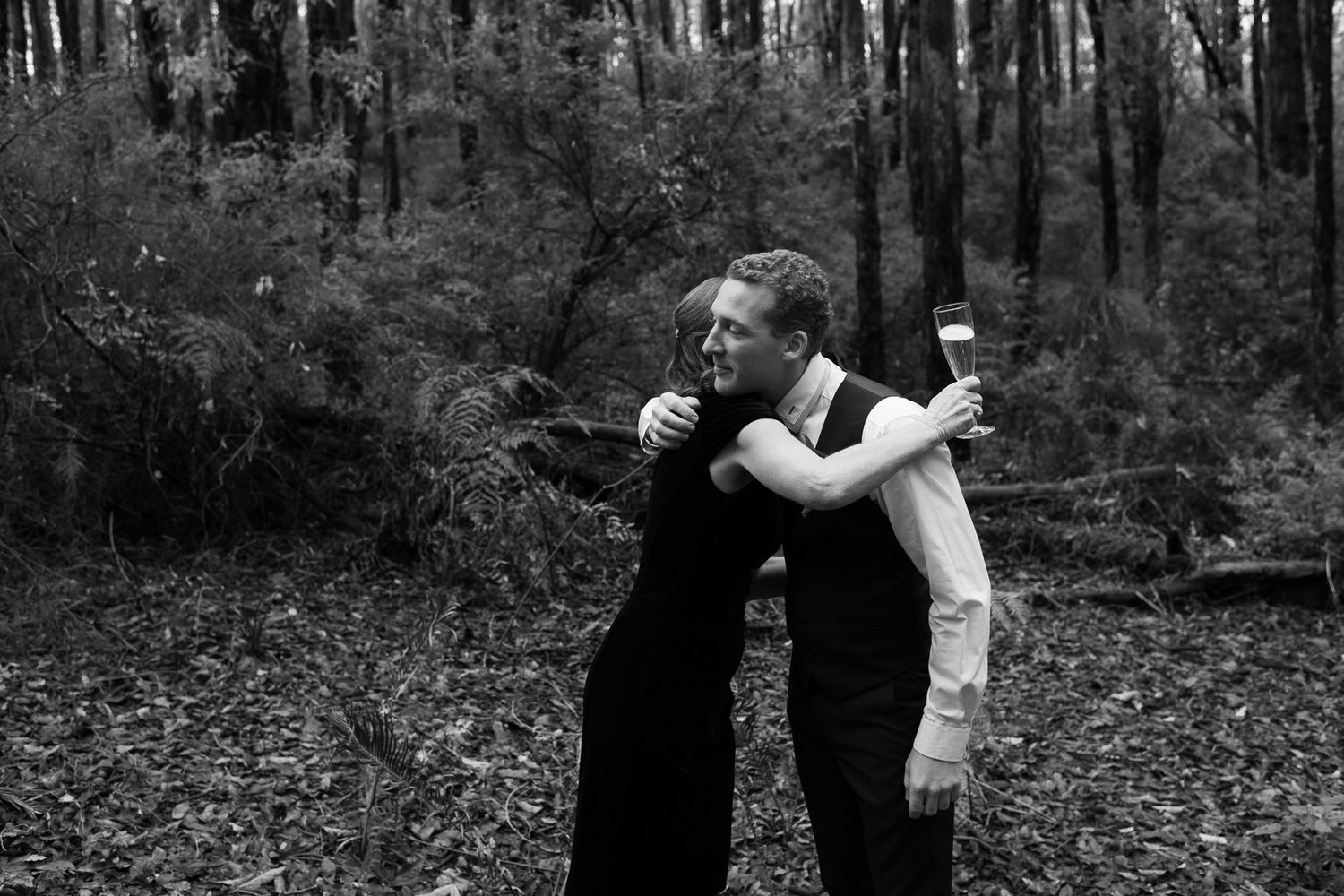 Two people embrace while dancing together in a dramatic black and white photo taken in a moody forest setting.