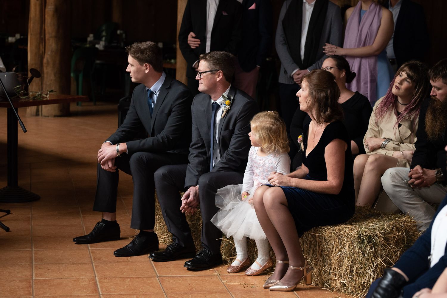 Wedding guests sit on hay bales during a rustic barn ceremony while a small child in white dress plays on the floor.