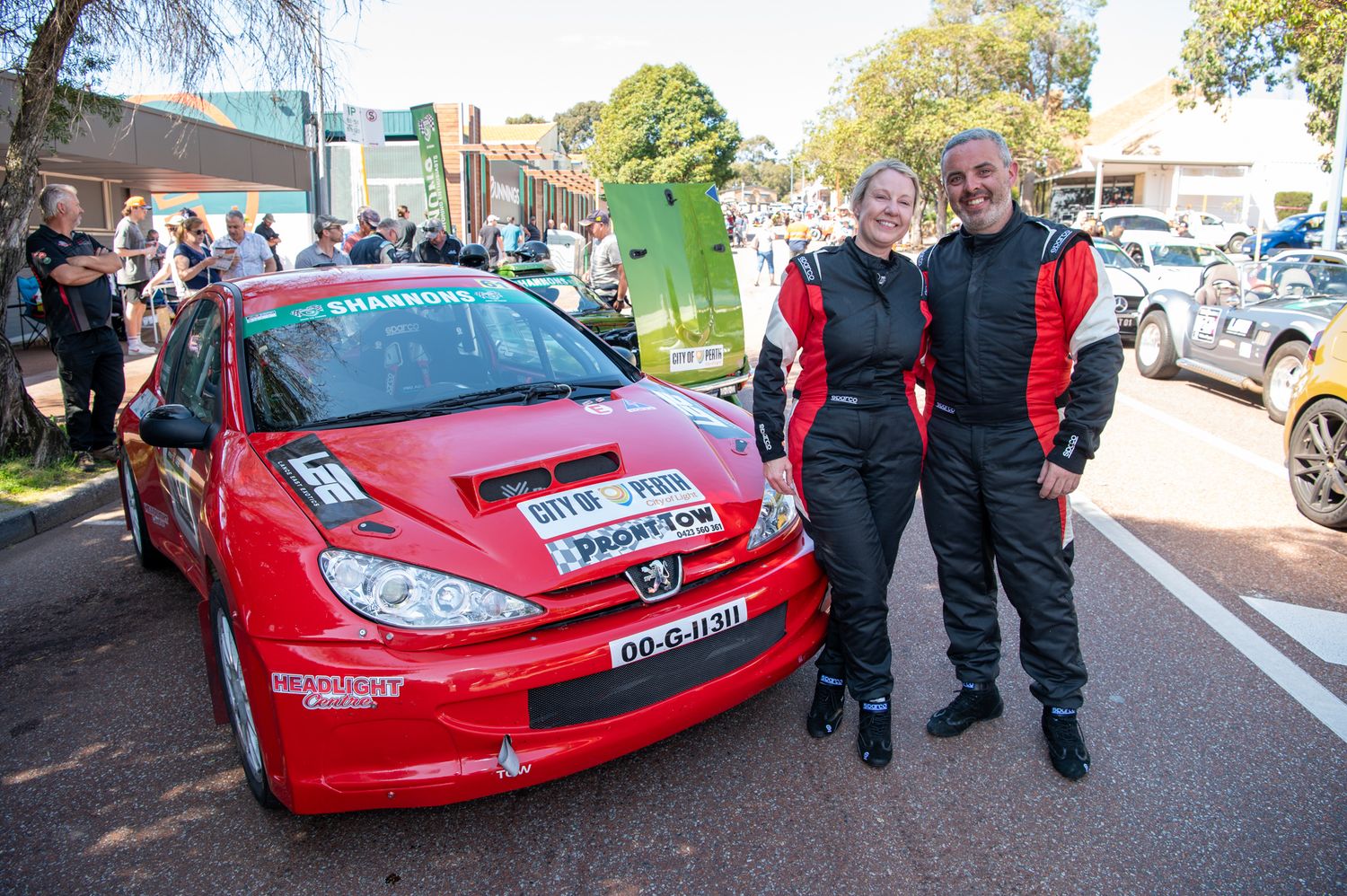 Red Peugeot rally car parked at motorsport event with competitors and spectators gathered around it.