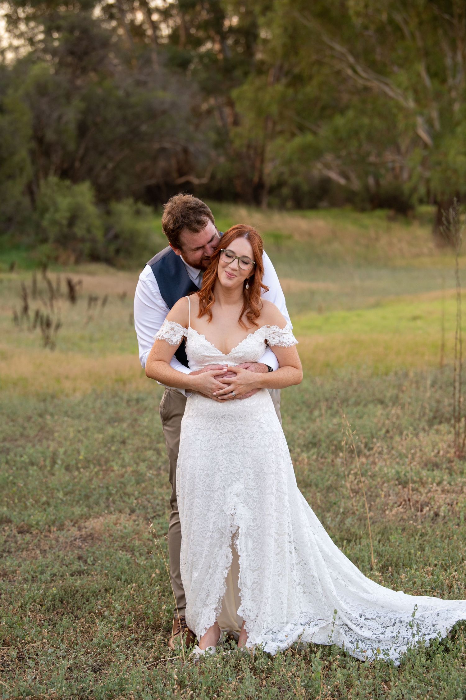 Romantic couple embracing on grassy hill during rustic outdoor wedding photoshoot.